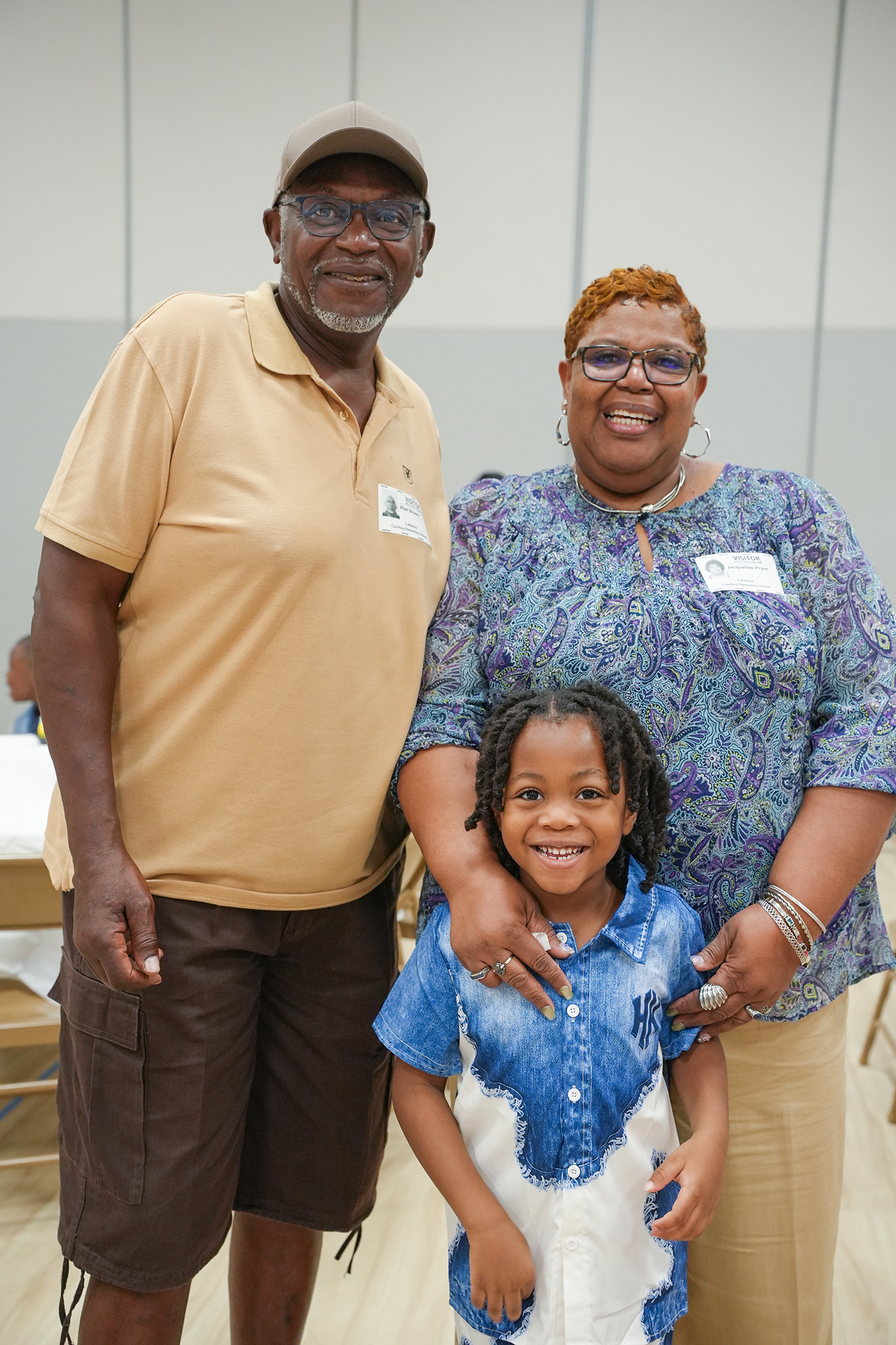 An adult man and woman with a young child, all smiling, standing in a room with chairs and tables.