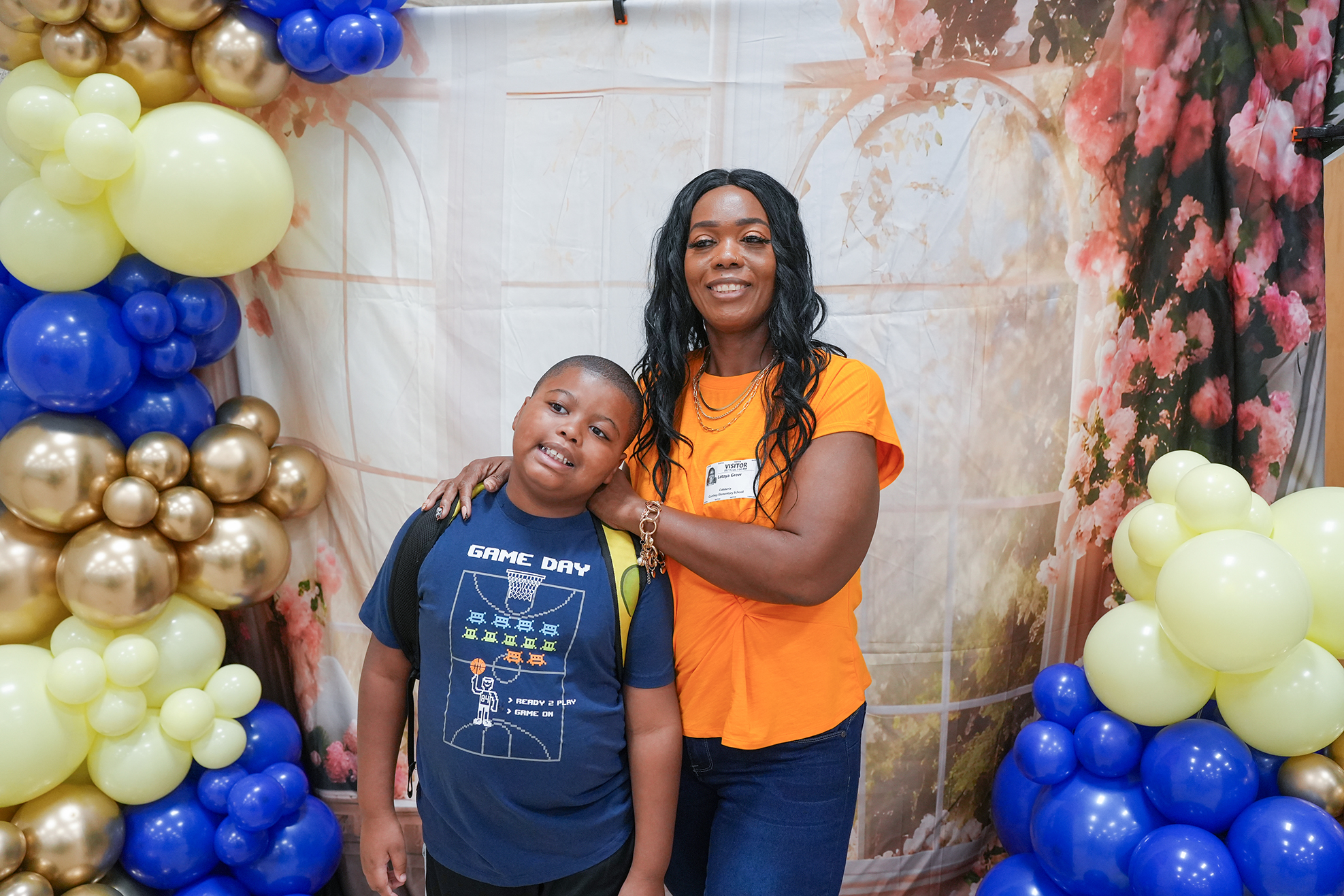 A woman in an orange shirt stands behind a smiling child in a blue shirt in a colorful balloon backdrop.
