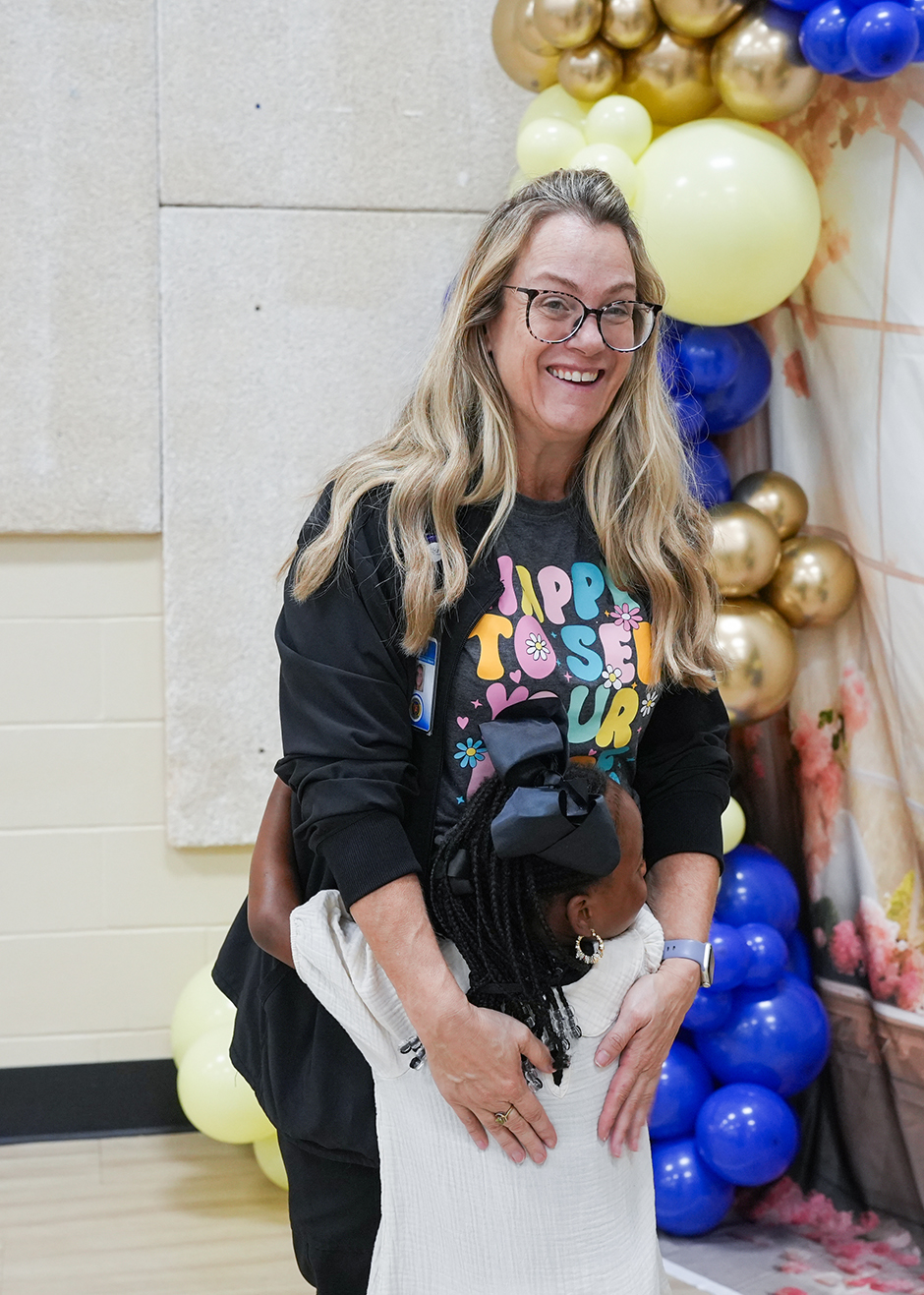 A woman wearing glasses and a black sweatshirt hugs a child wearing a white dress. They stand before balloons.