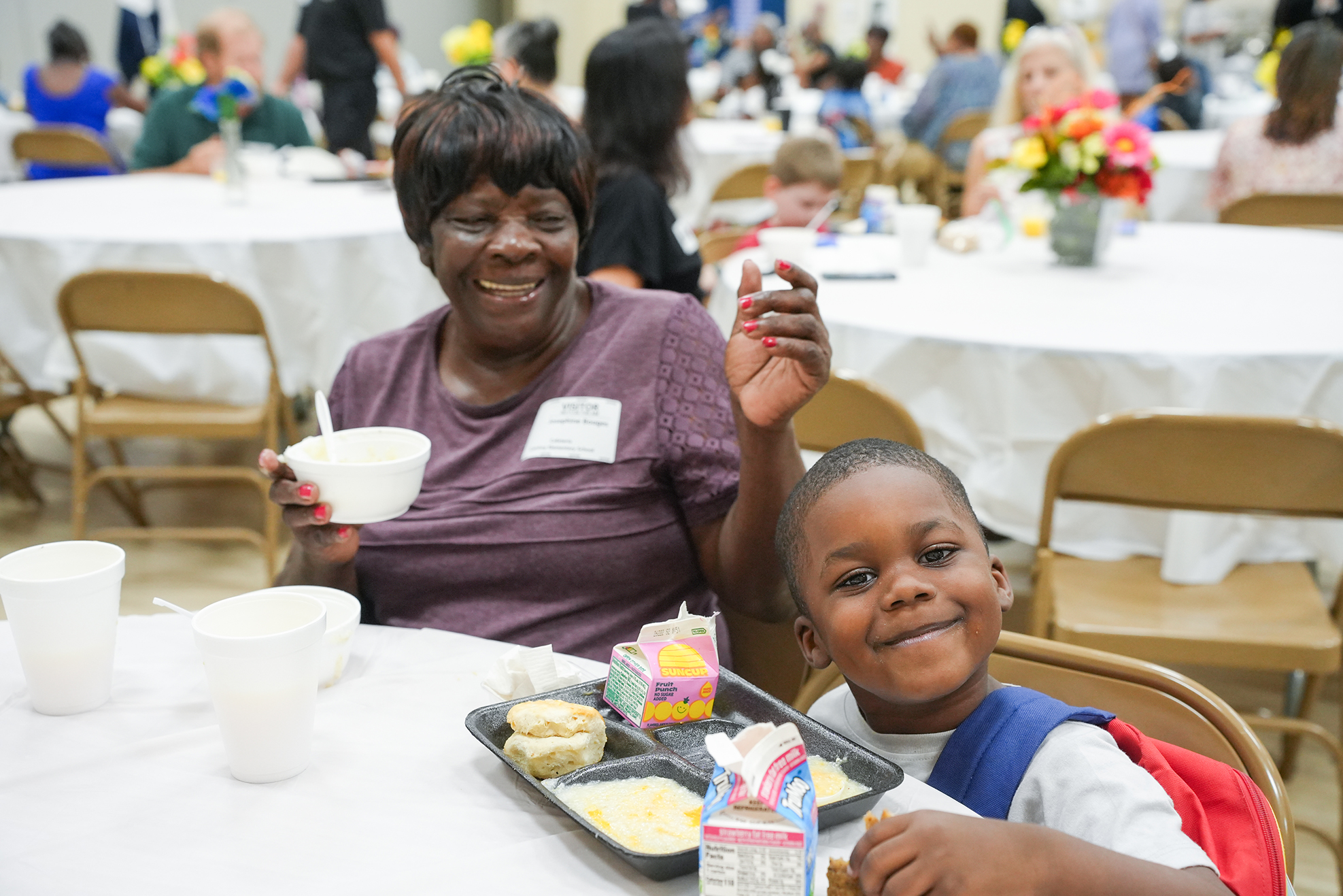 An adult and child seated at a table, smiling. Trays of food, cups, and a flower arrangement.