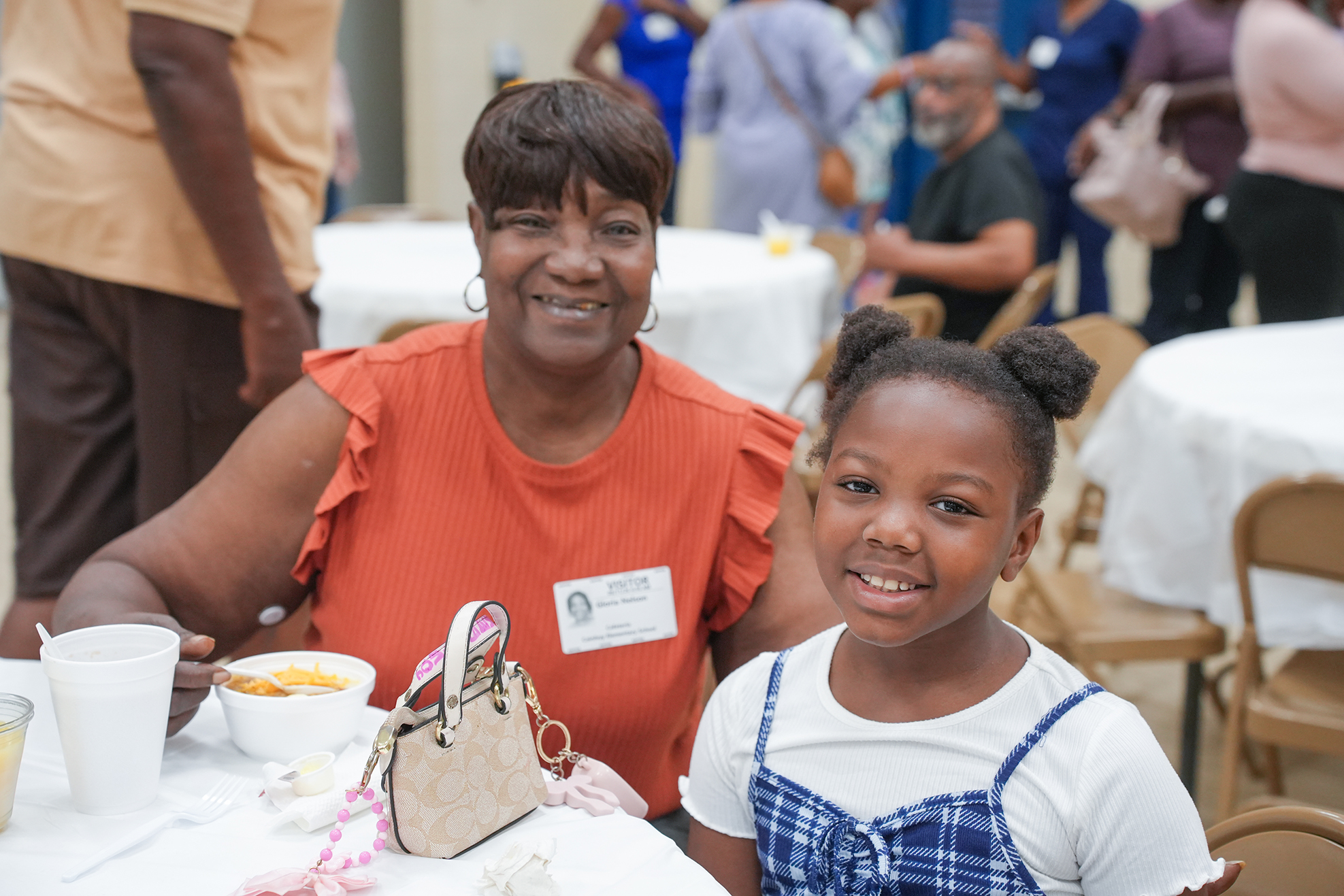 A woman in an orange shirt with a girl child, both smiling, at a table with cups.
