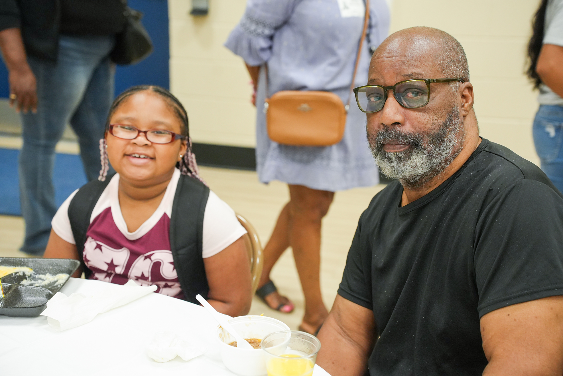 A man and a girl sit at a table. The girl has a backpack and glasses. A woman with a handbag walks behind them.