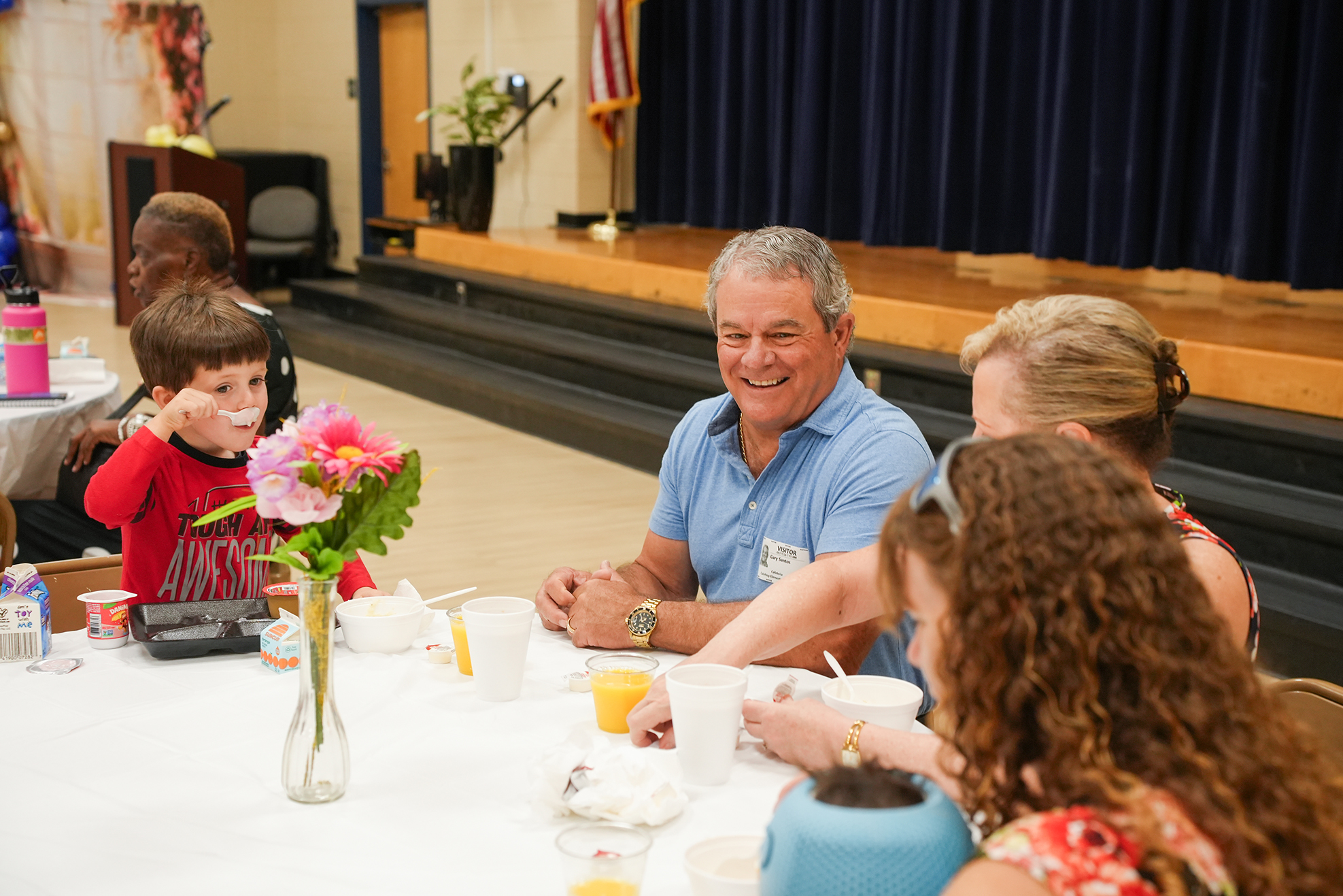 A group of adults and children seated around a table with flowers, cups, and bowls, smiling.