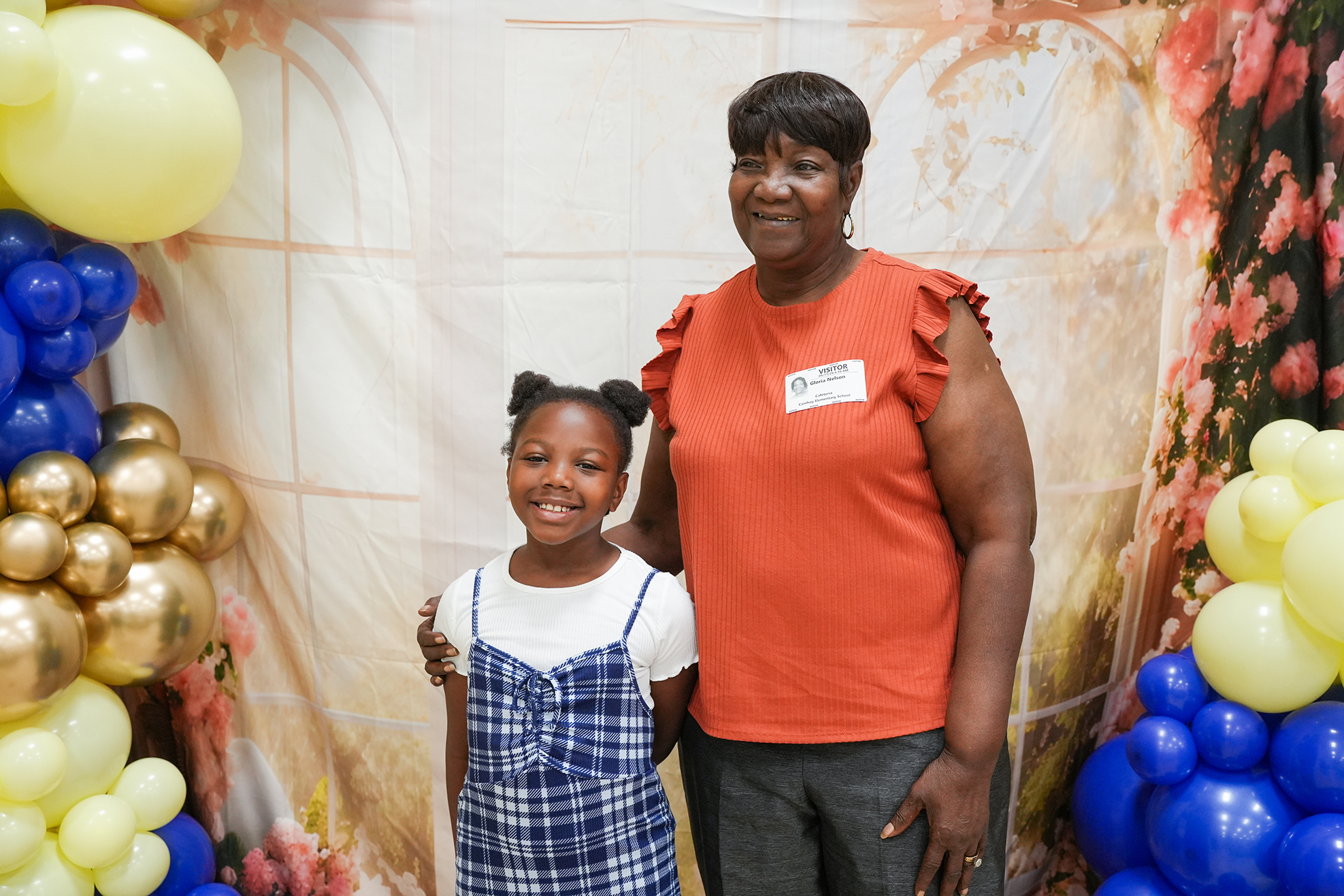 A woman and a girl stand together in front of balloons. The woman wears an orange shirt. The girl wears a white dress.