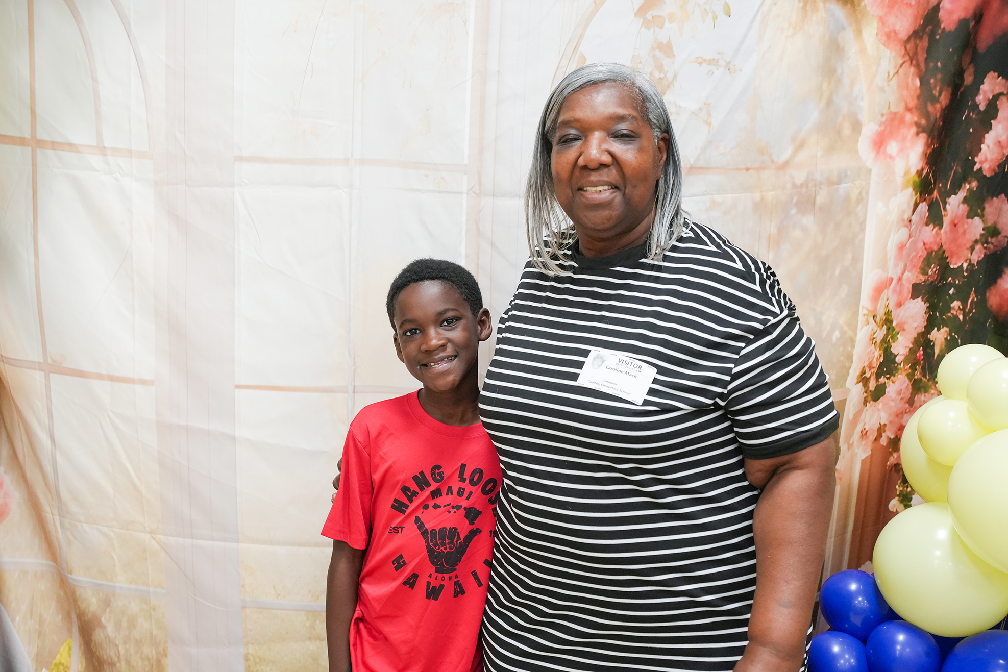 A smiling woman with gray hair stands beside a boy in a red t-shirt. Behind them is a white backdrop with a floral design and colorful balloons.