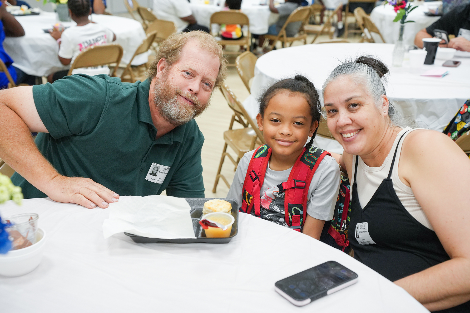 A man and a woman sit at a table with a child between them. They smile at the camera.