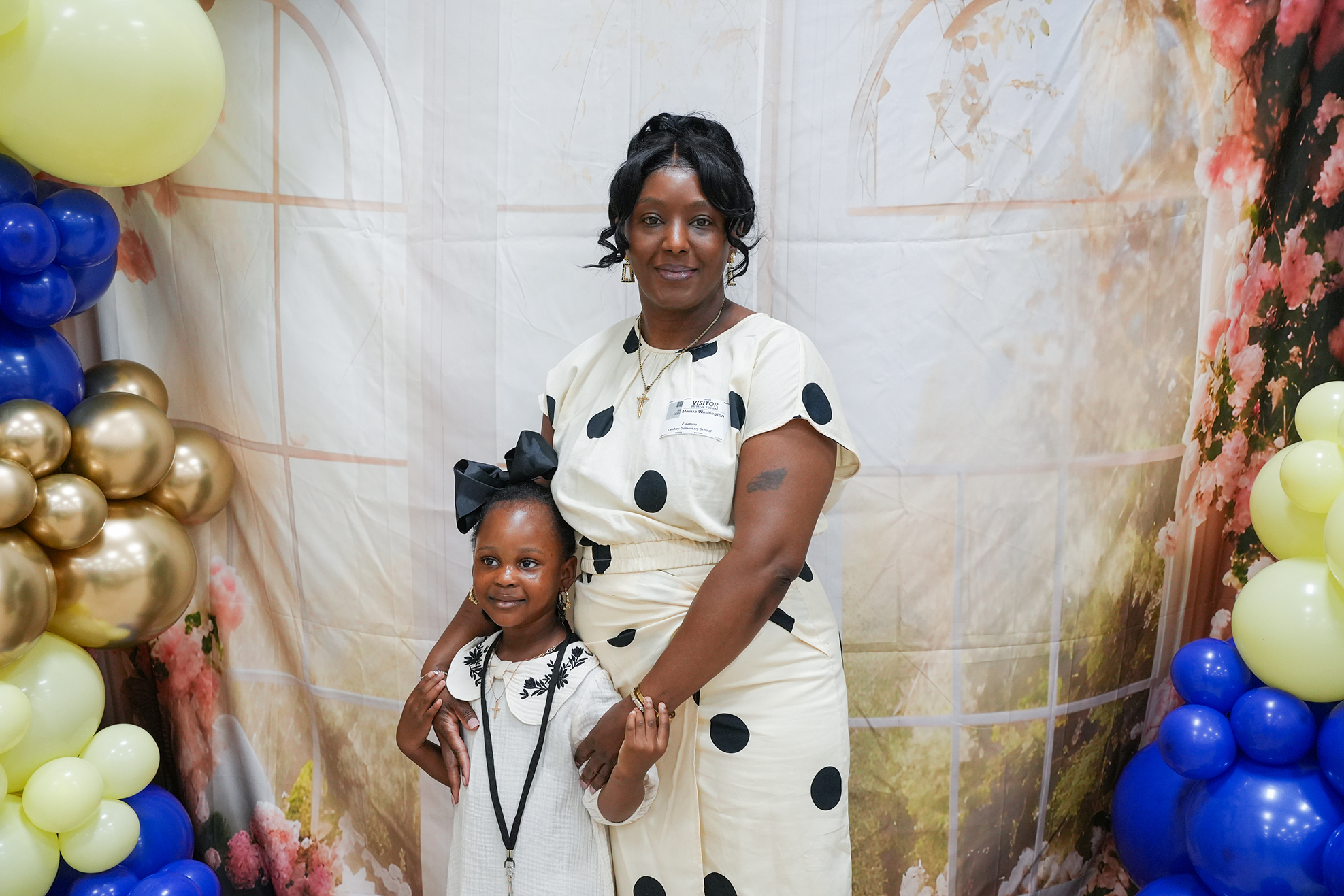A woman and a child stand before a backdrop of flowers and balloons. The woman wears a polka-dot dress.