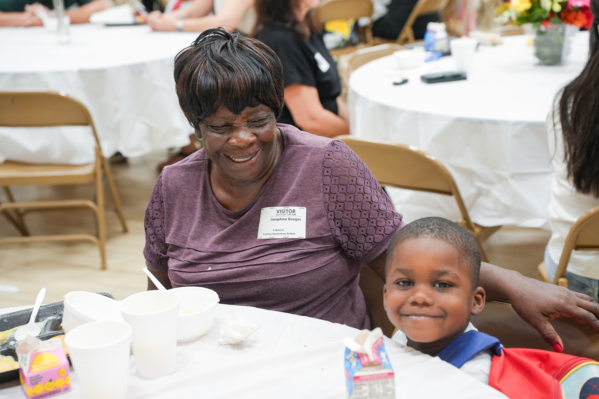 Woman and child sitting at a table, smiling, with food and drink. Behind them, tables with white cloths and chairs.