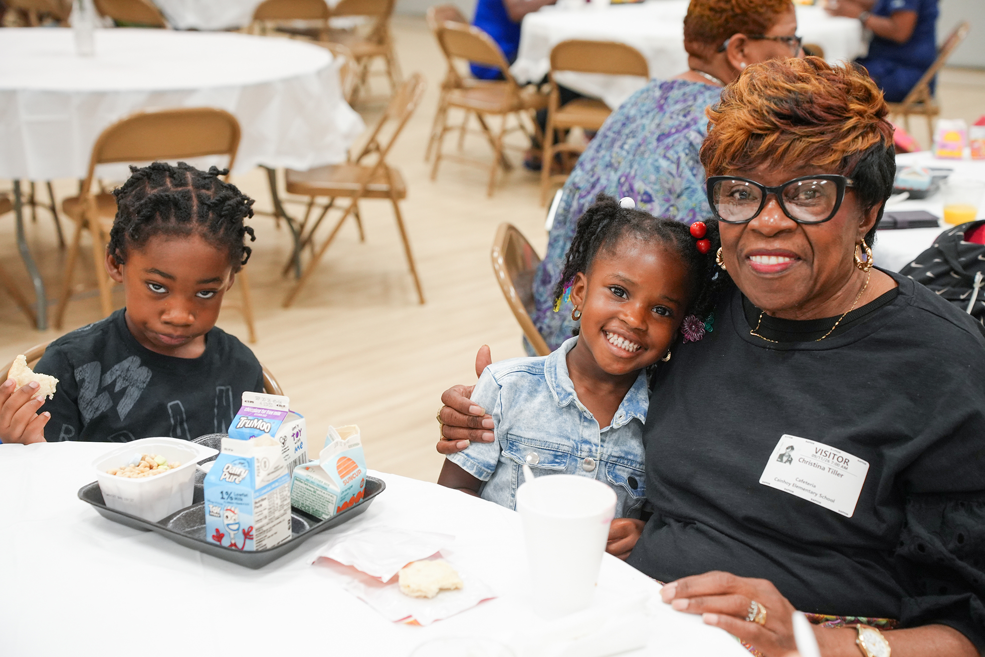 An adult woman with short hair and glasses holds two young girls at a table. One girl smiles, and the other eats.