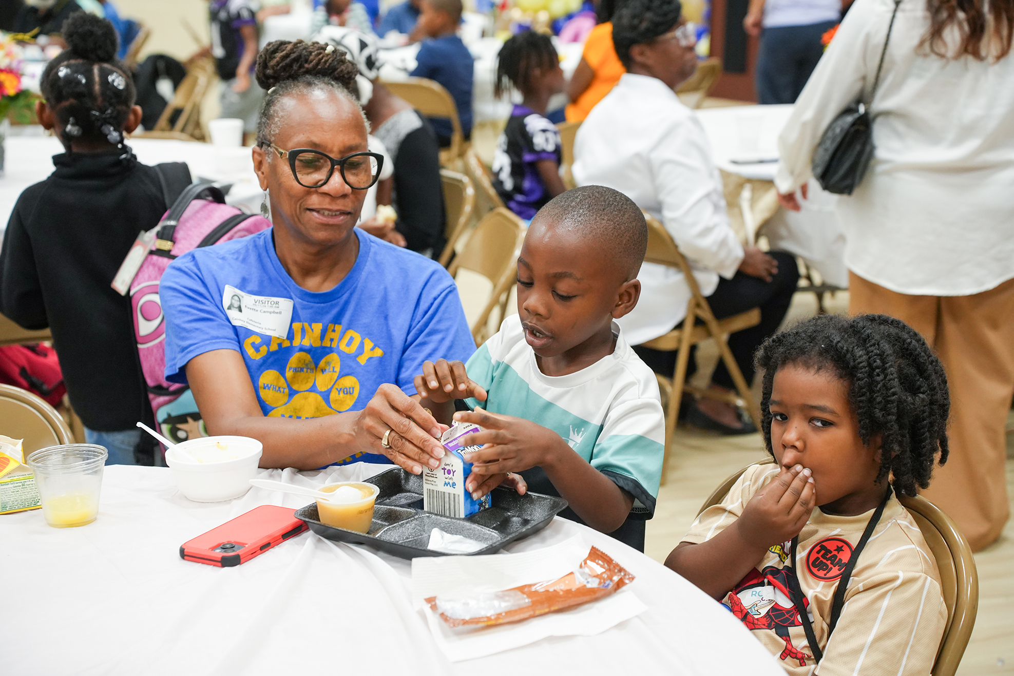 Woman and two kids sit at a table. Boy holds a carton. Another child eats. People in the background.