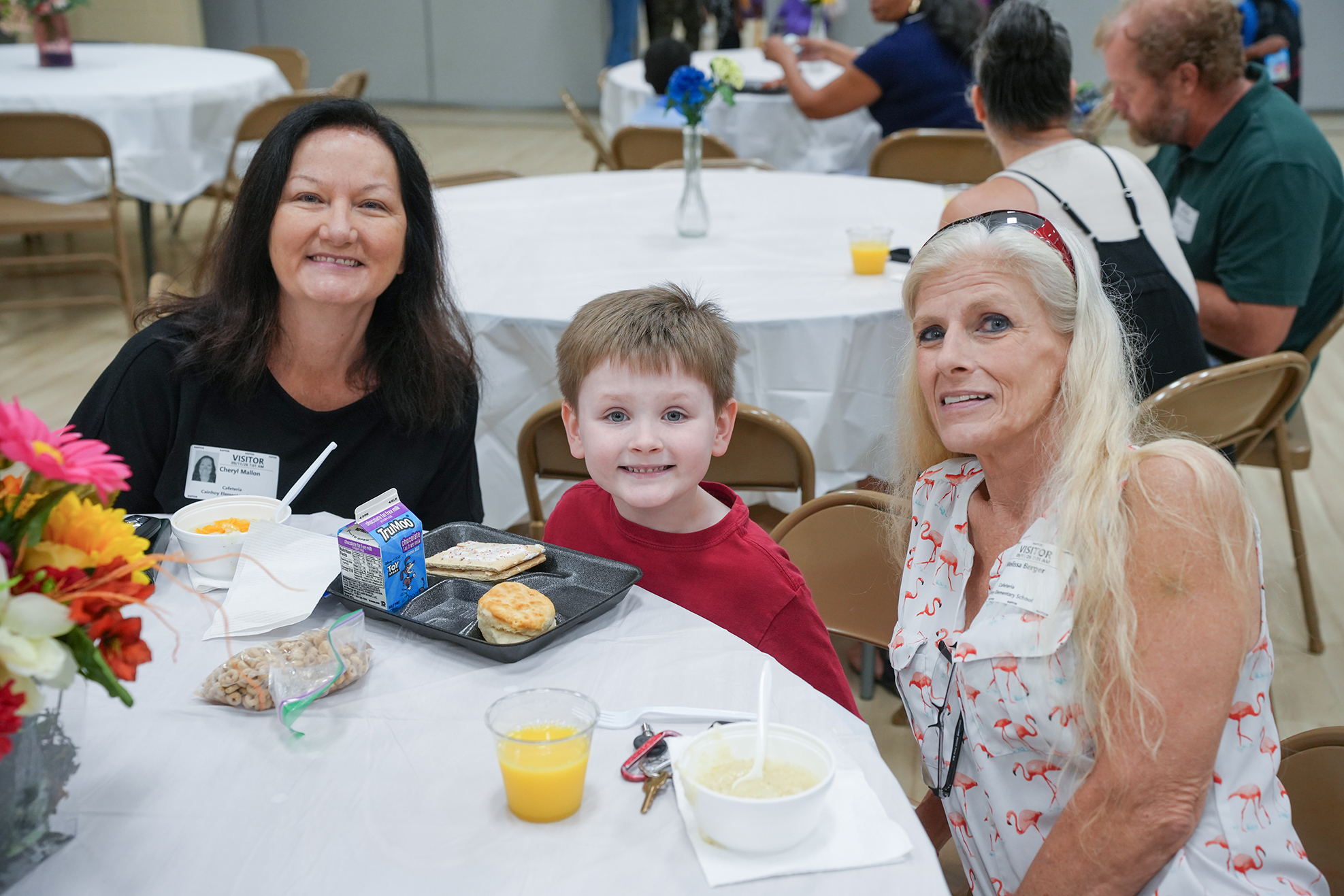 Three people at a table with food. A child in a red shirt, a woman in black, and a woman with blonde hair.