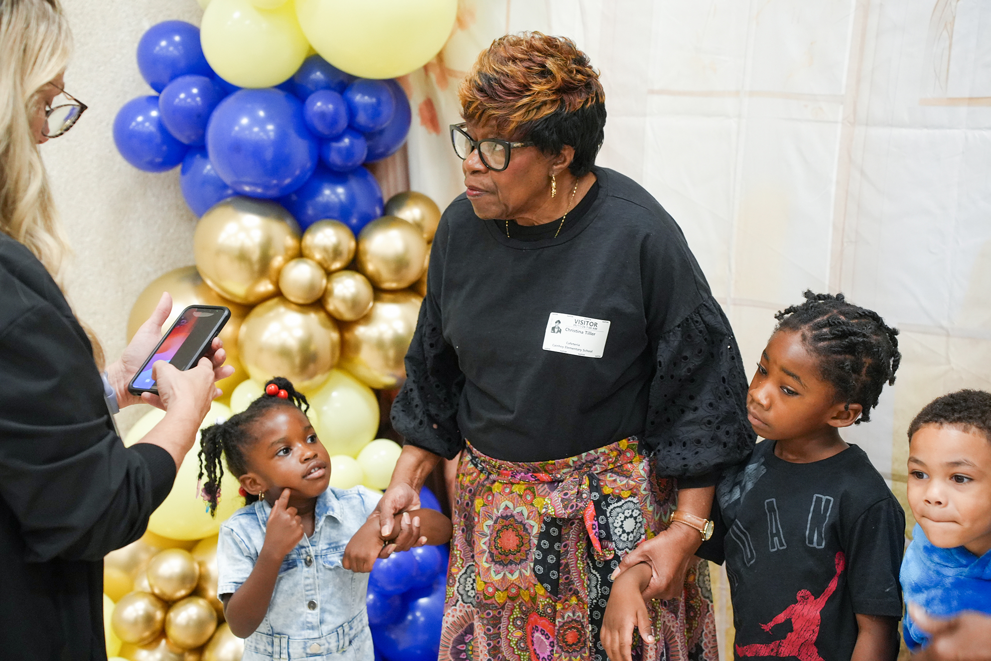 A woman speaks to a group of children near a backdrop of multicolored balloons. One child holds a cell phone.