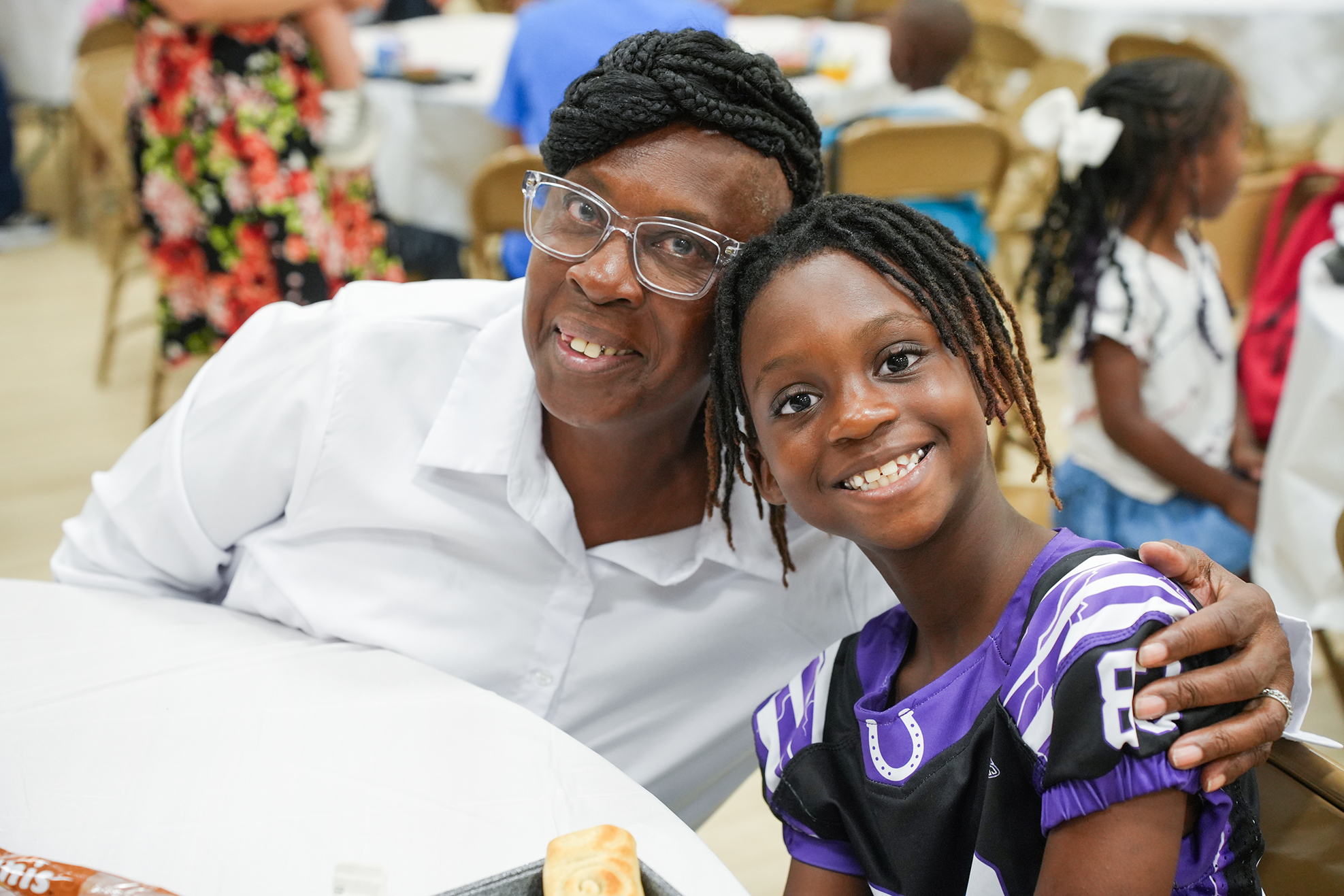 An adult and a child sit together at a table. The child has their arm on the adult's shoulder.
