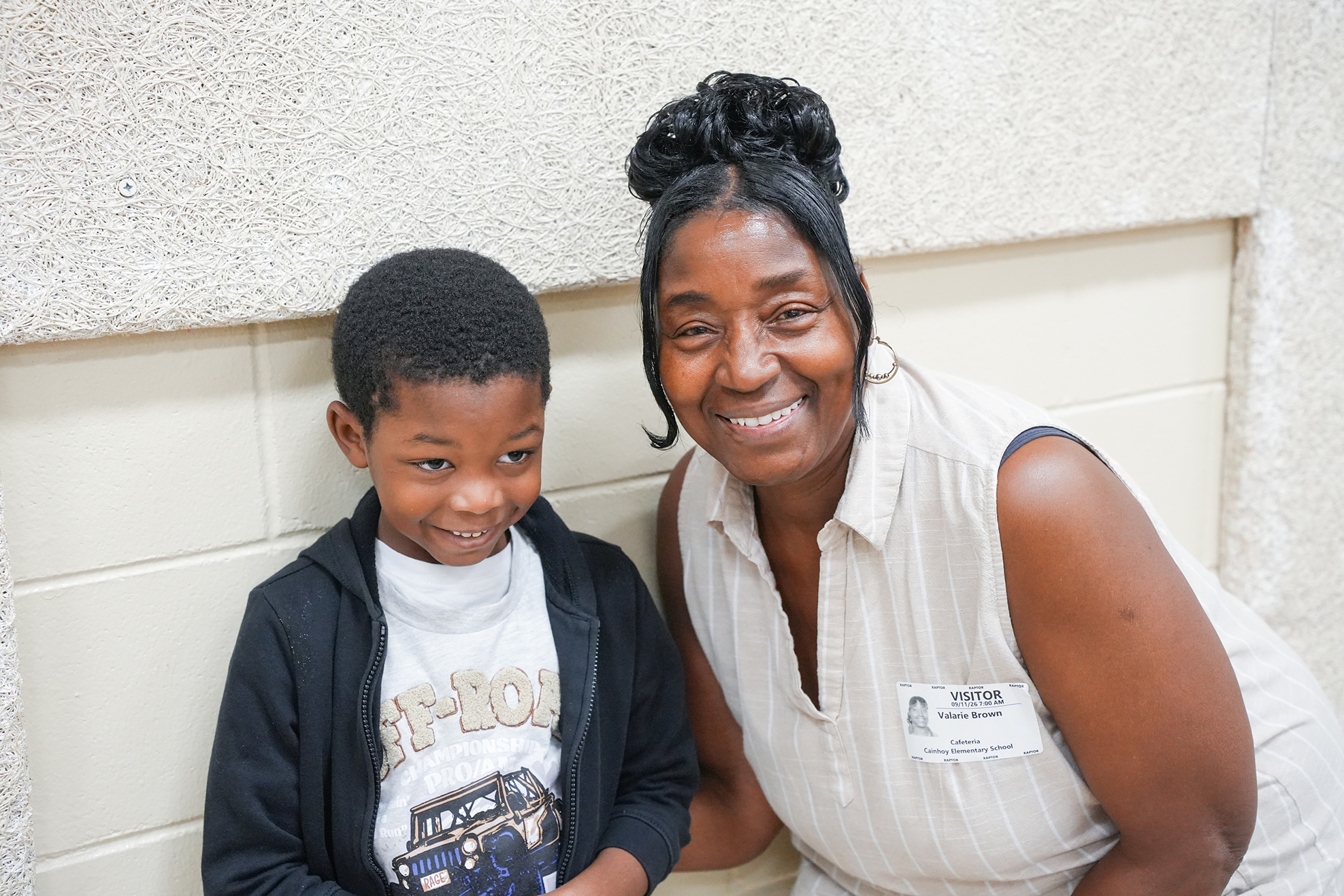 A woman and a boy sit next to each other, both smiling. They sit against a tiled wall.