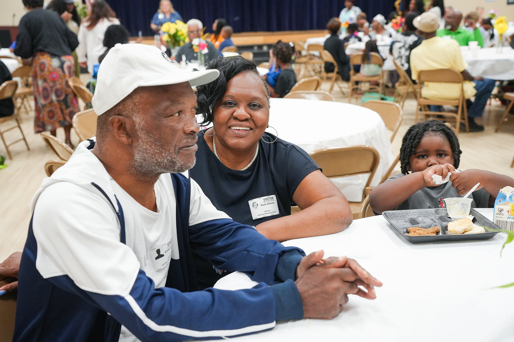 An older man, woman, and child sit at a table with trays of food in a dining room.