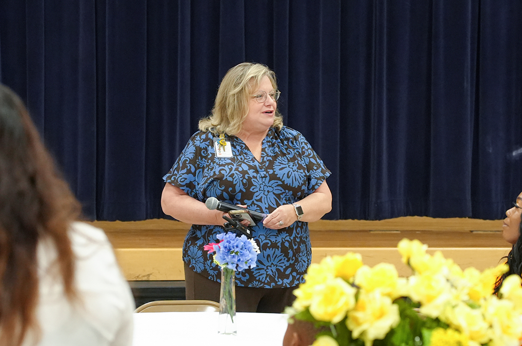 A woman speaks into a microphone, wearing a blue shirt with a flower design. She stands in front of a table with a flower arrangement.