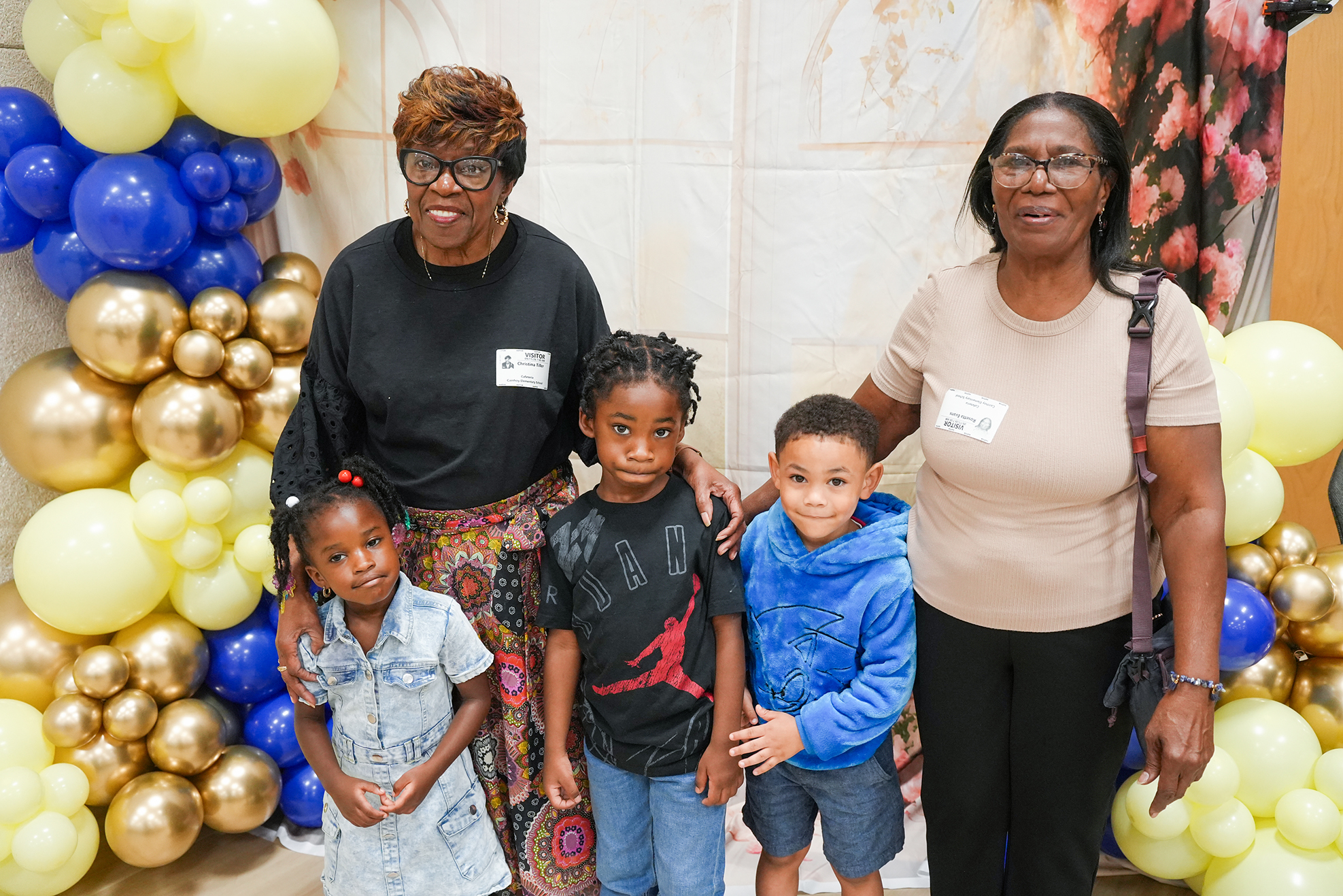 Group of women and children pose in front of a backdrop with balloons in gold, blue, and yellow.