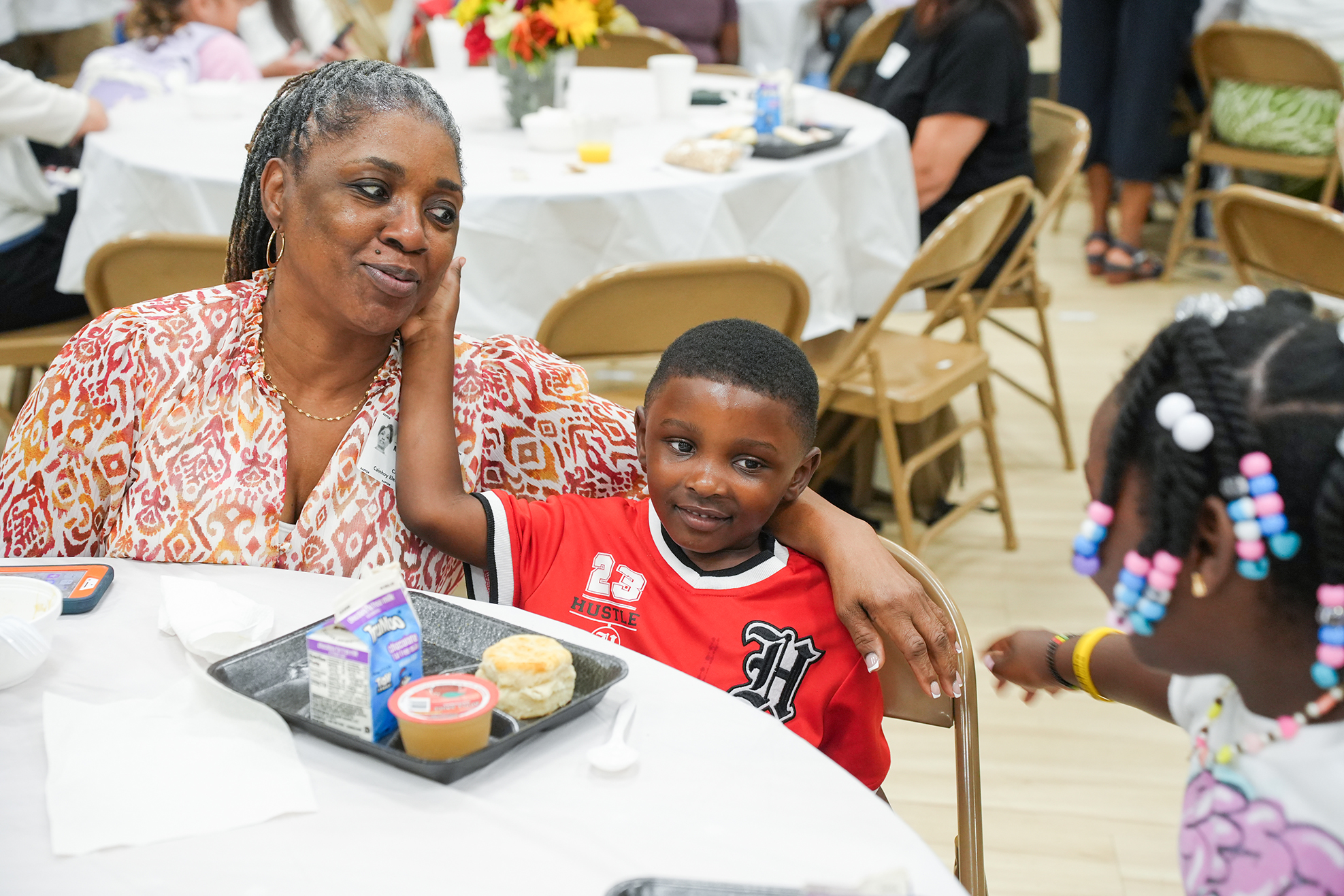 A woman and two children at a table. The child in the red shirt is smiling at the camera.