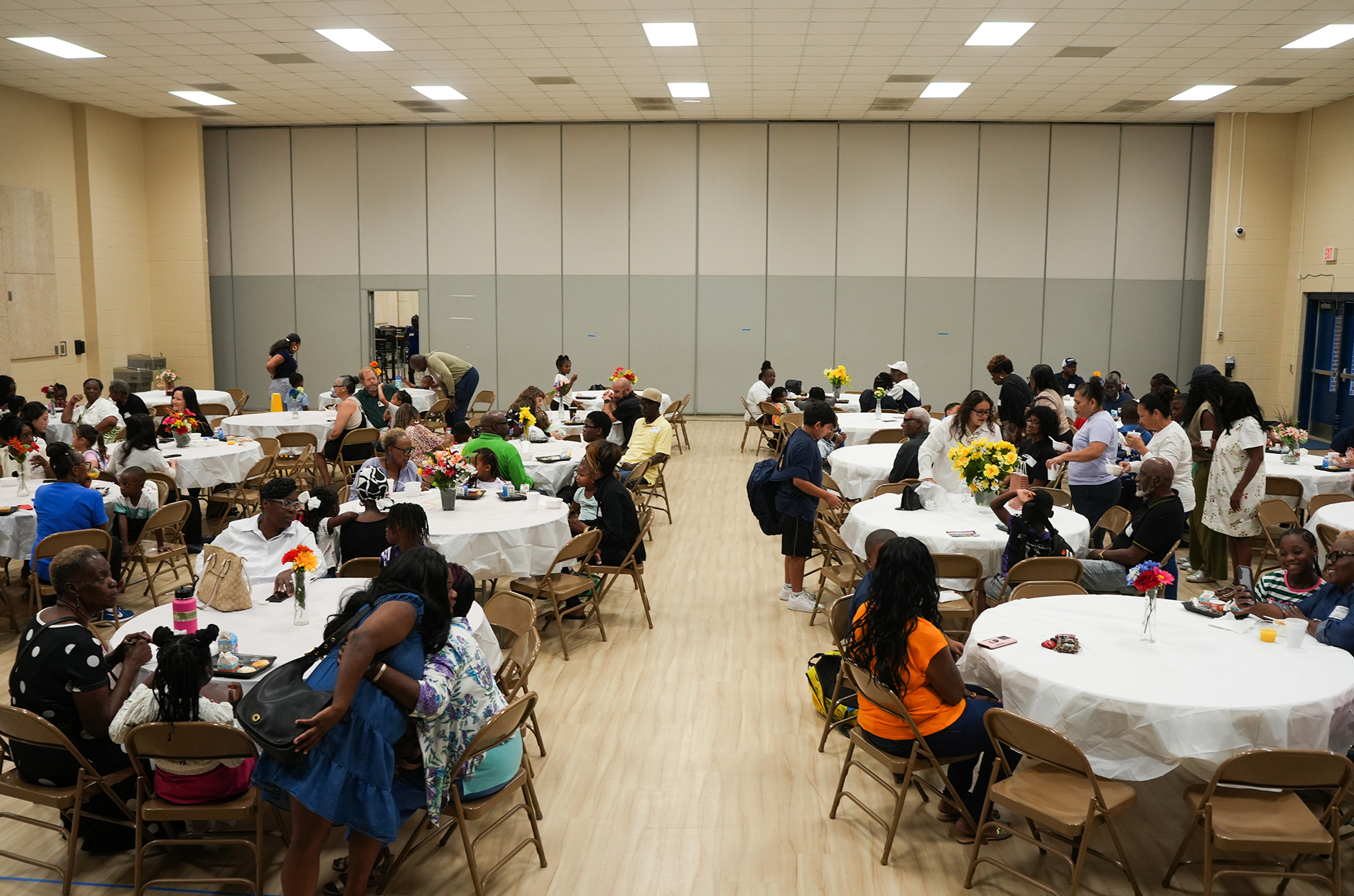 A dining hall with tables and chairs. Tables have white tablecloths and flowers. Attendees sit, stand, and move around.
