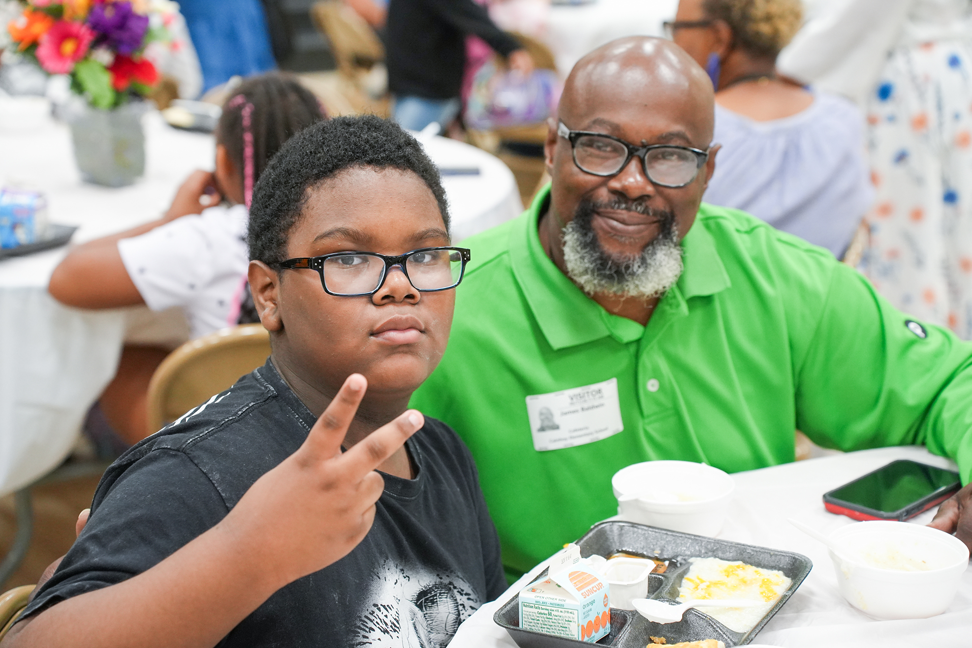 A boy wearing glasses sits at a table making a peace sign. Next to him is a man with a beard and glasses.