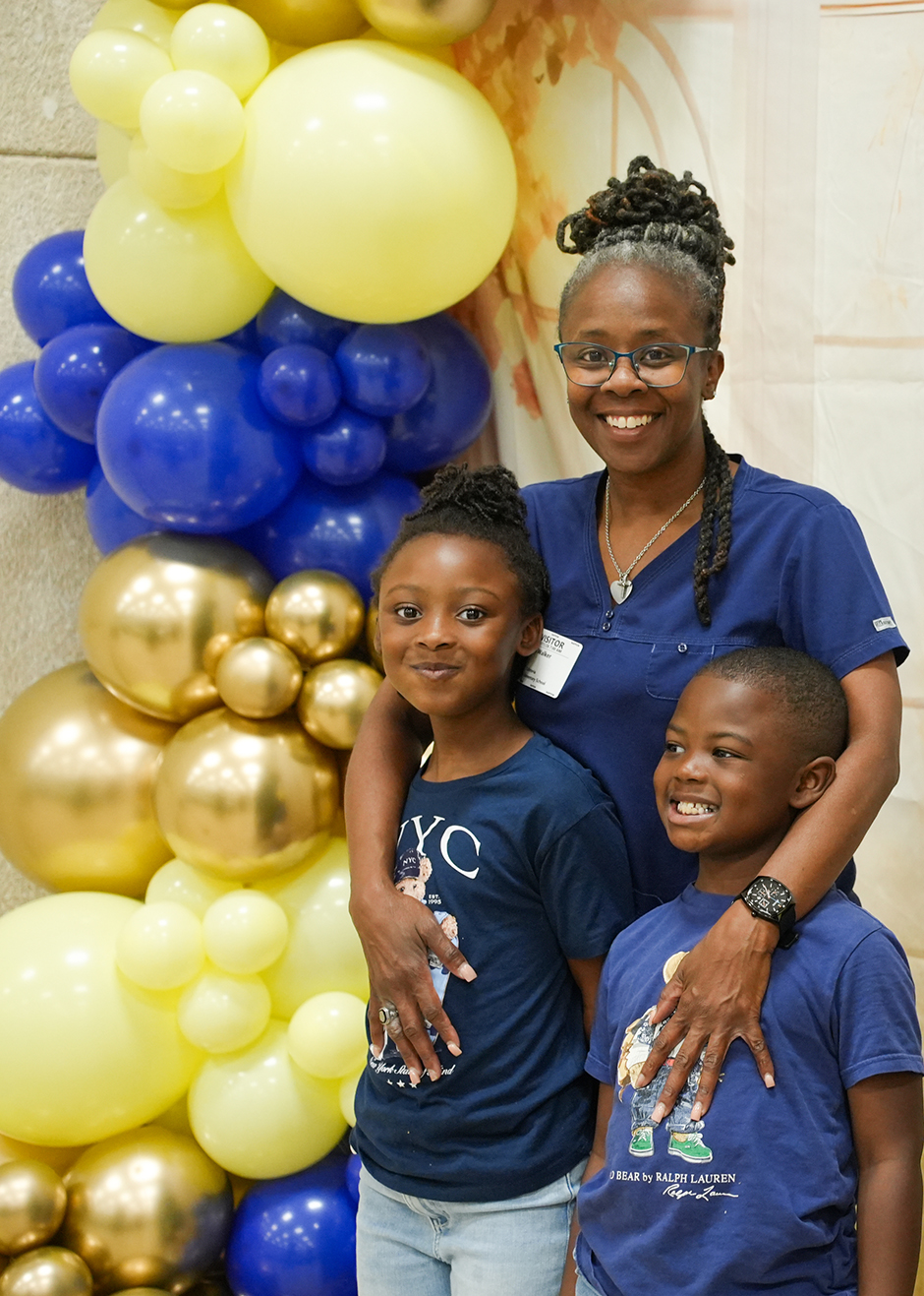 A woman in blue and two children in blue pose in front of a colorful balloon arch.