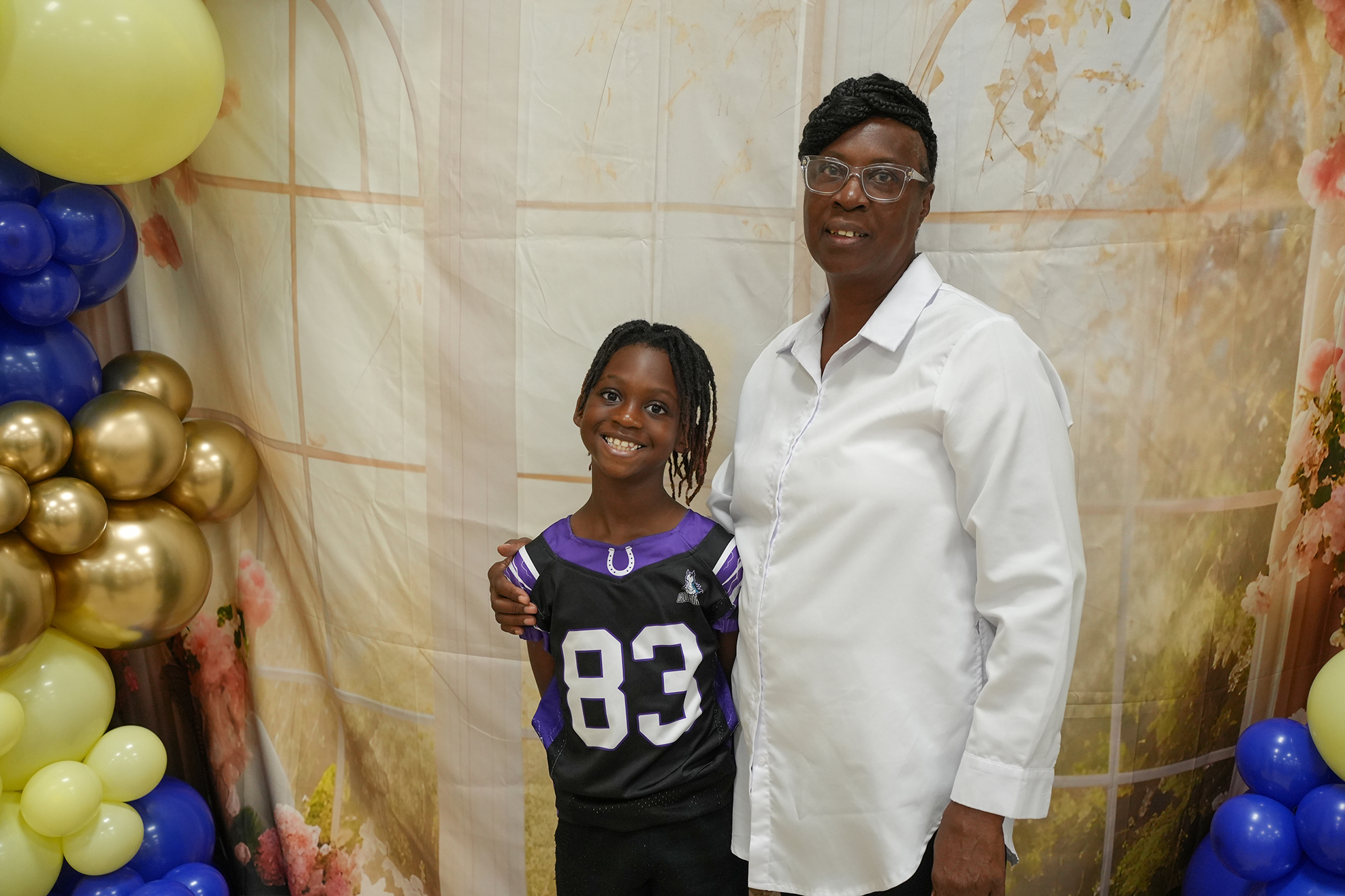 A woman and a young girl stand together against a backdrop with balloons. The woman wears glasses, and the girl wears a black and purple sports jersey.