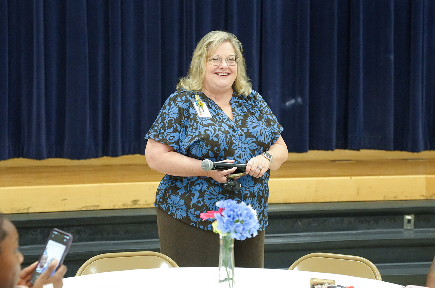 A woman with a microphone stands in front of a stage with a closed curtain. She is surrounded by a table with a blue flower vase.