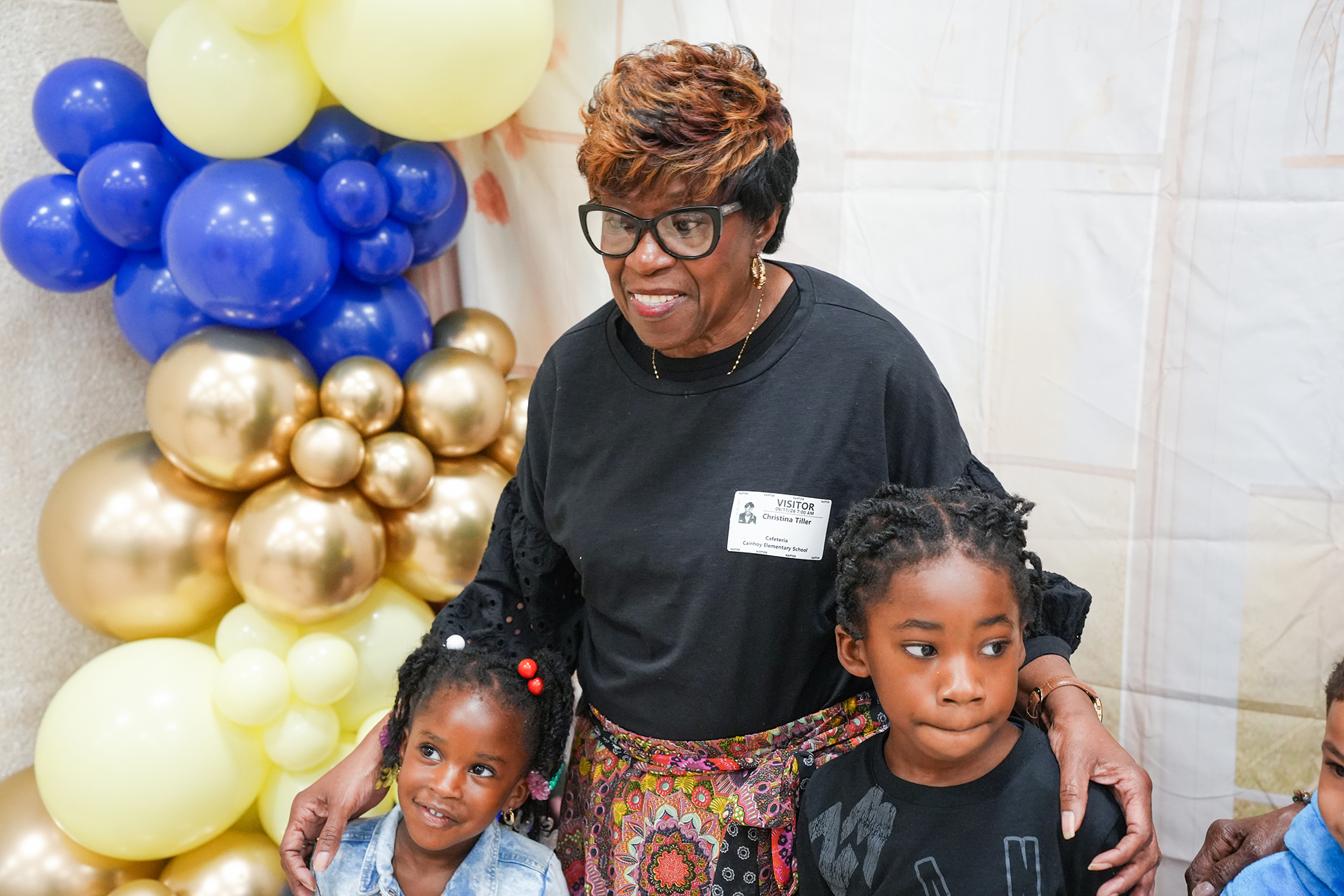 Woman with two girls under a large balloon arch. They all have name tags and are smiling.