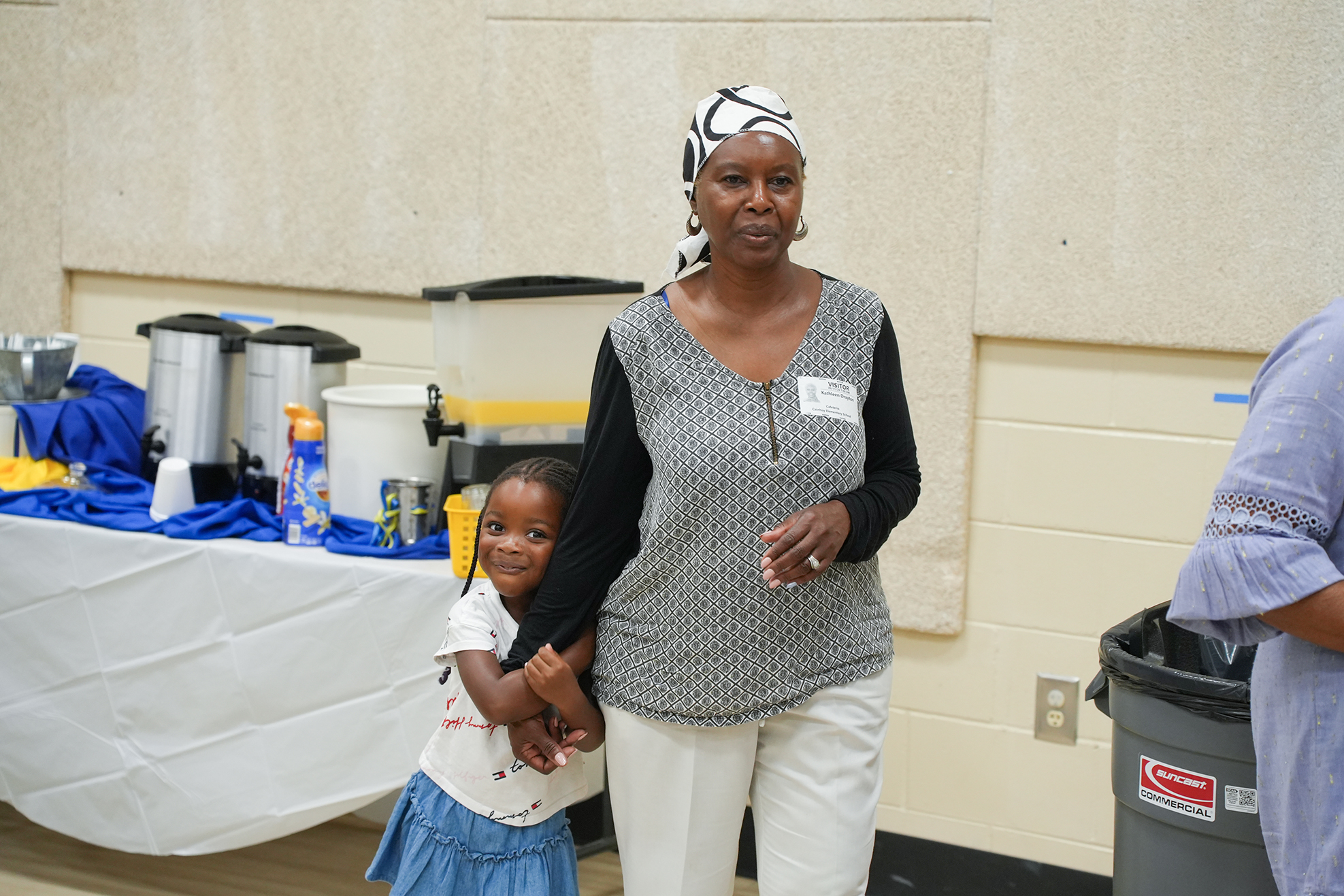 An adult and child stand together in a room with a table, trash bin, and a beige wall.