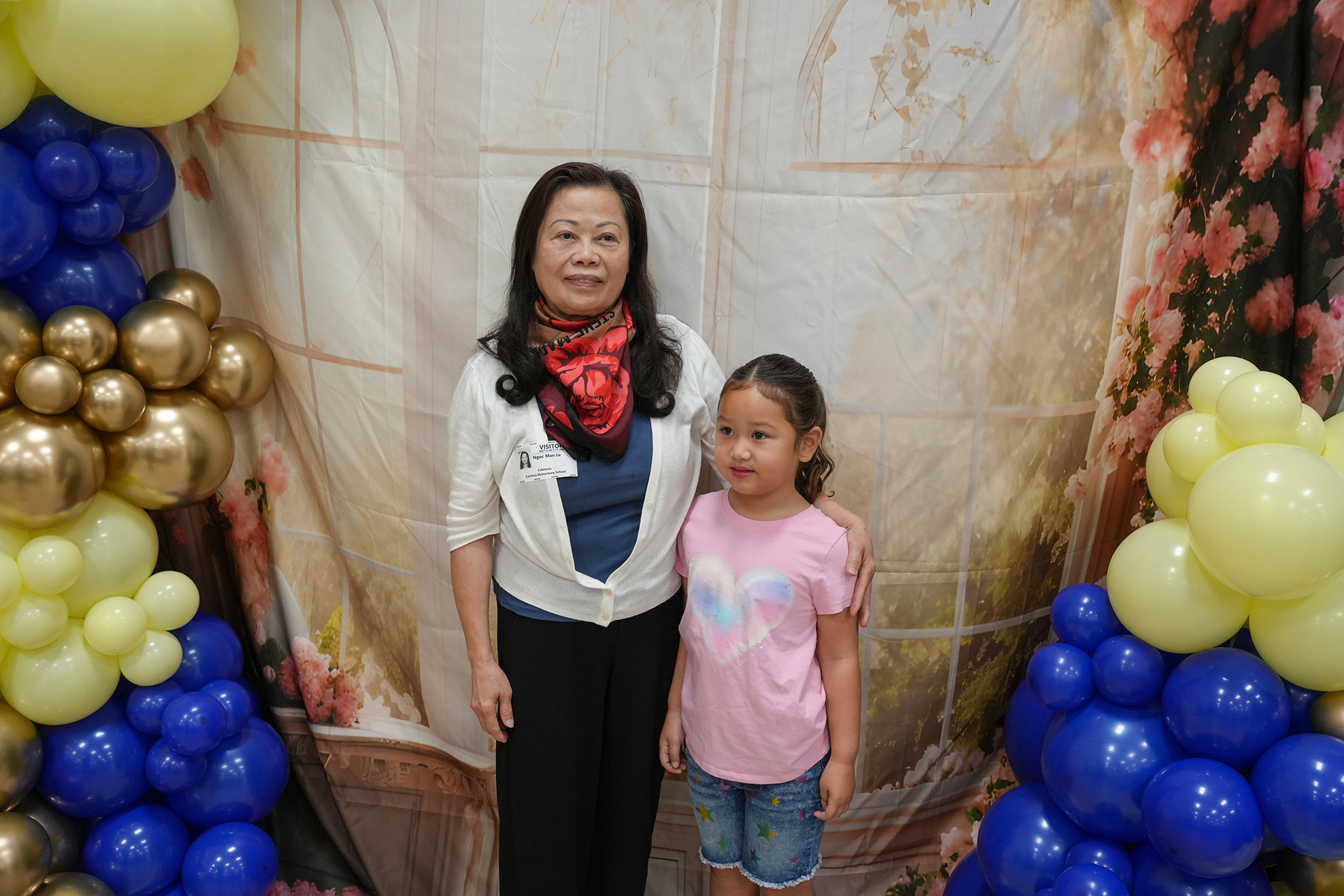 Adult and child in front of a balloon arch, posing for a photo. Background features a floral and window design.