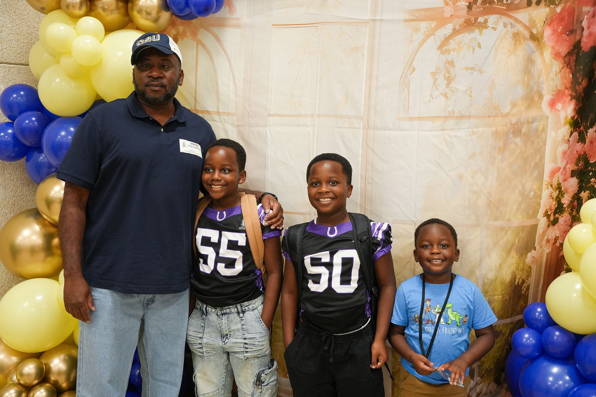 An adult man and three children stand together, smiling. Behind them, a backdrop with balloons.