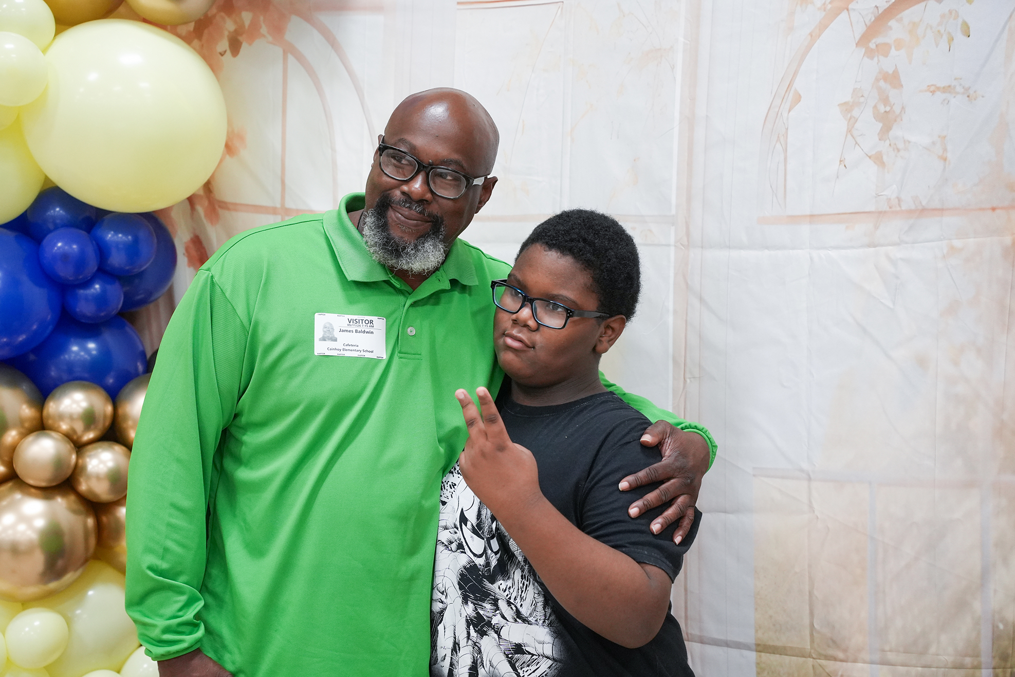Man with a green shirt and eyeglasses embraces a boy in a black shirt at a photo booth.