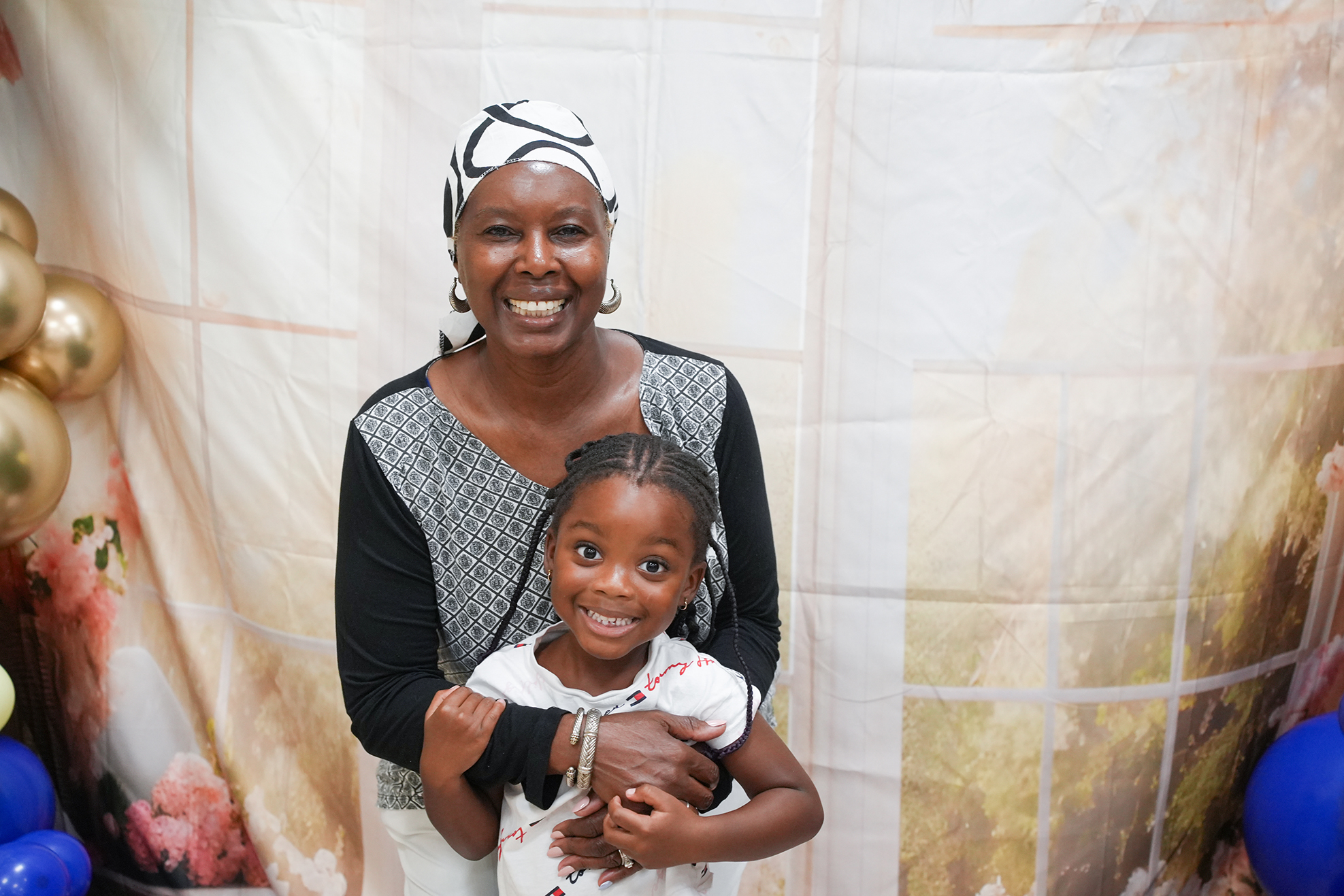 Woman in a white scarf holding a girl with pigtails and a white dress in front of a backdrop.