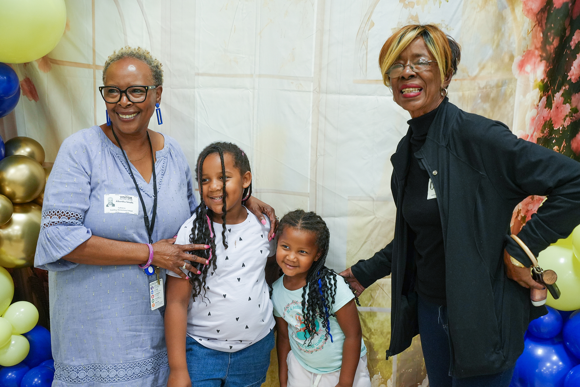 Three women and two young girls stand smiling near a backdrop with balloons. The women wear glasses and accessories, while the girls wear casual clothing.