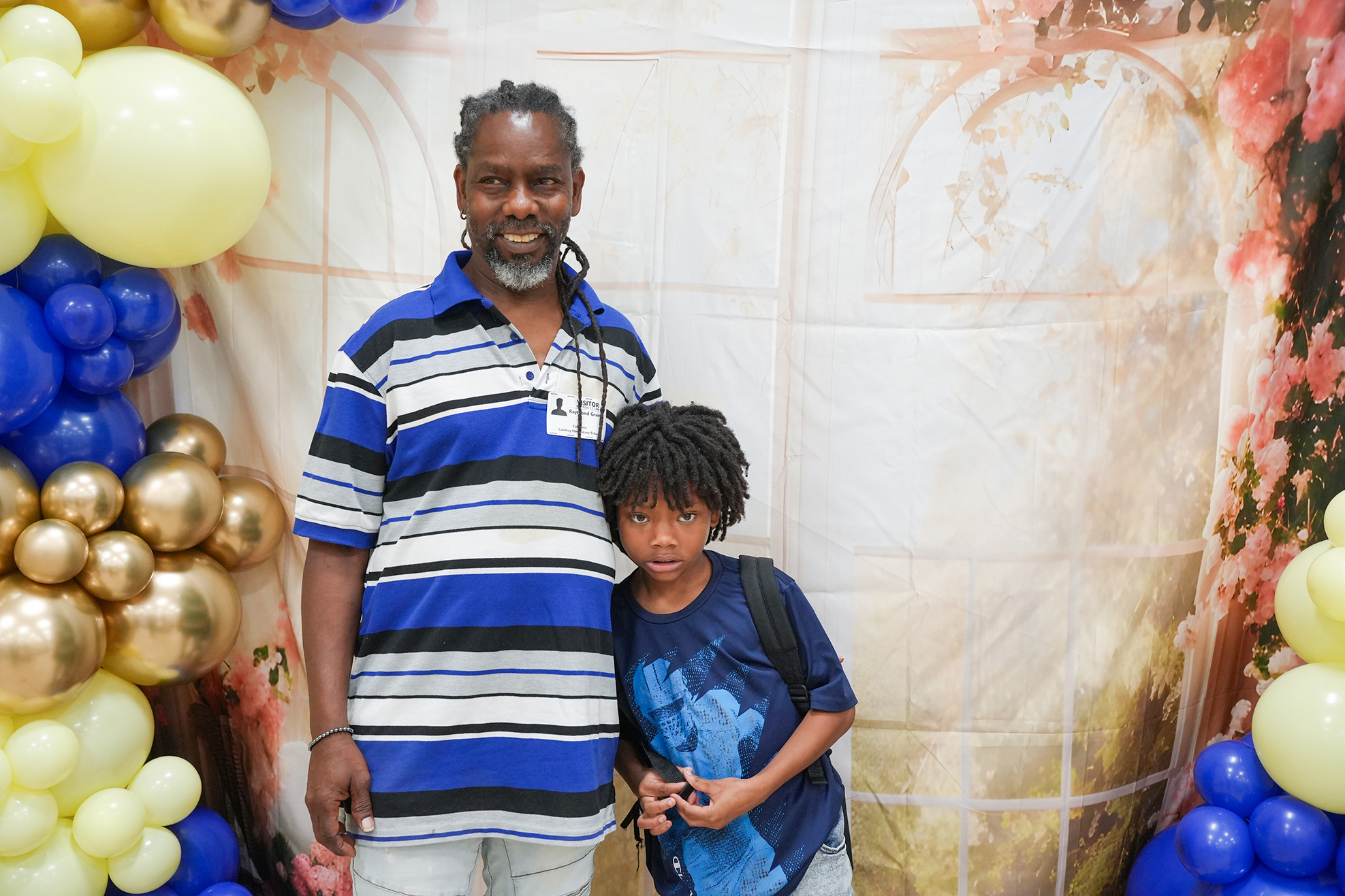A man and a boy stand before balloons, with the man wearing a blue and white striped shirt.