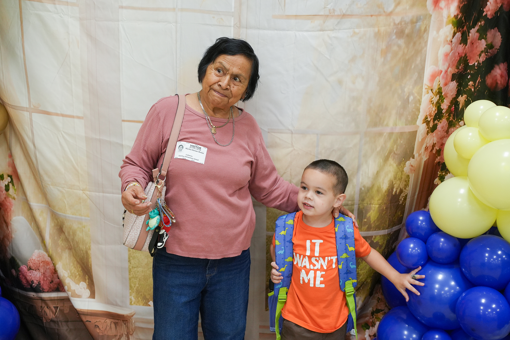 A woman with a child at an event. She holds a bag and stands behind a child in orange.