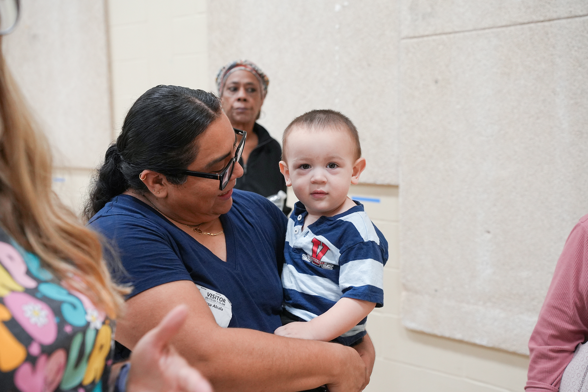 A woman holds a baby in a room. Other people are in the background. The walls are white.