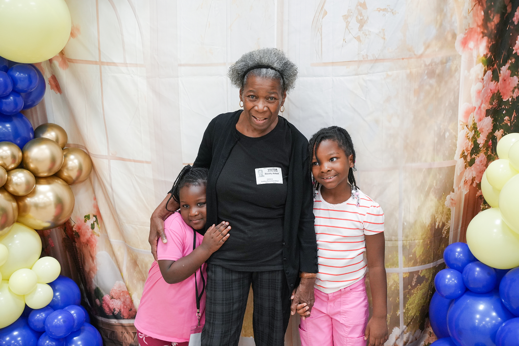 An adult and two children standing behind a backdrop with balloons, smiling for a picture.
