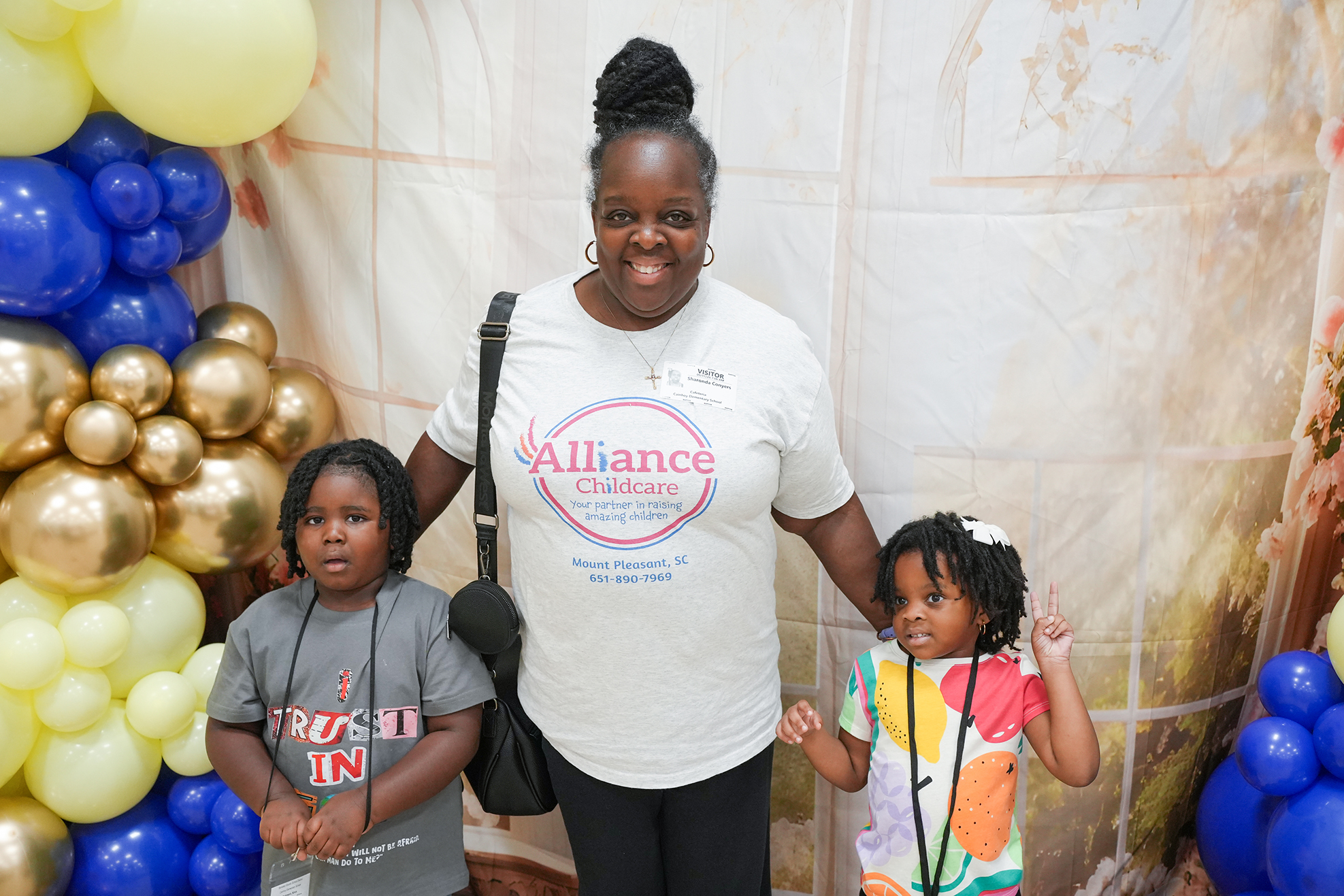 A woman and two children stand in front of a colorful backdrop with balloons. The woman wears a white shirt.