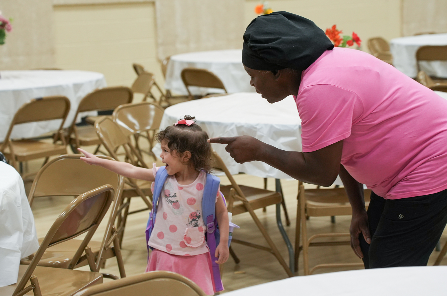 Woman pointing at a young girl with a backpack, with tables and chairs in the background.