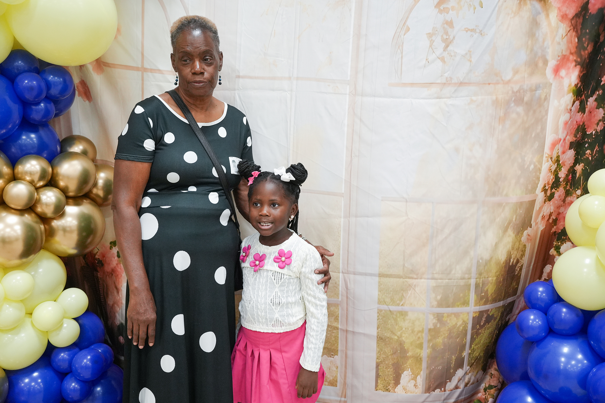 A woman and a young girl stand near balloons. The woman wears a polka-dot dress, and the girl wears a white shirt and pink skirt.