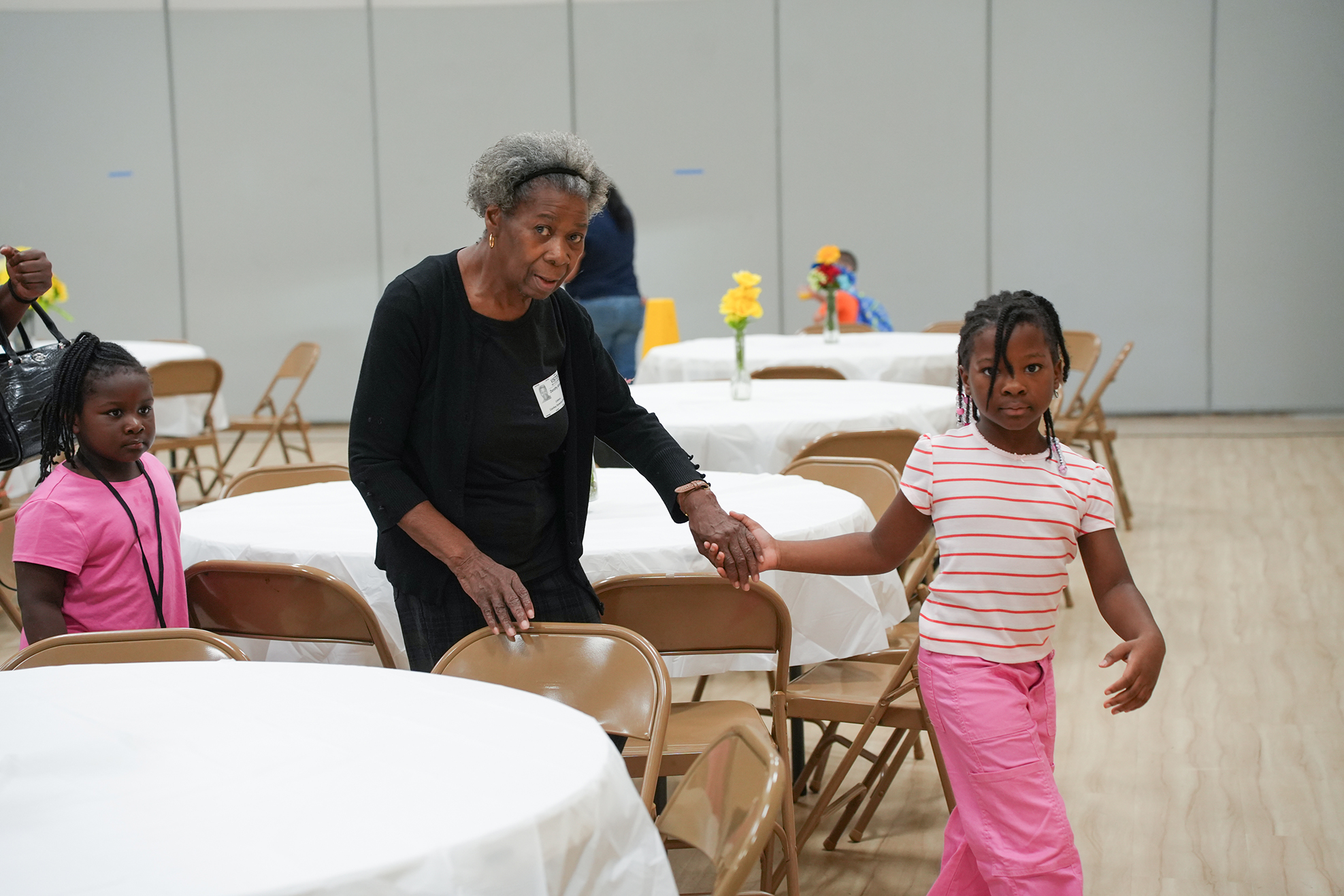 An adult holds hands with a child. They are in a room with tables and chairs.