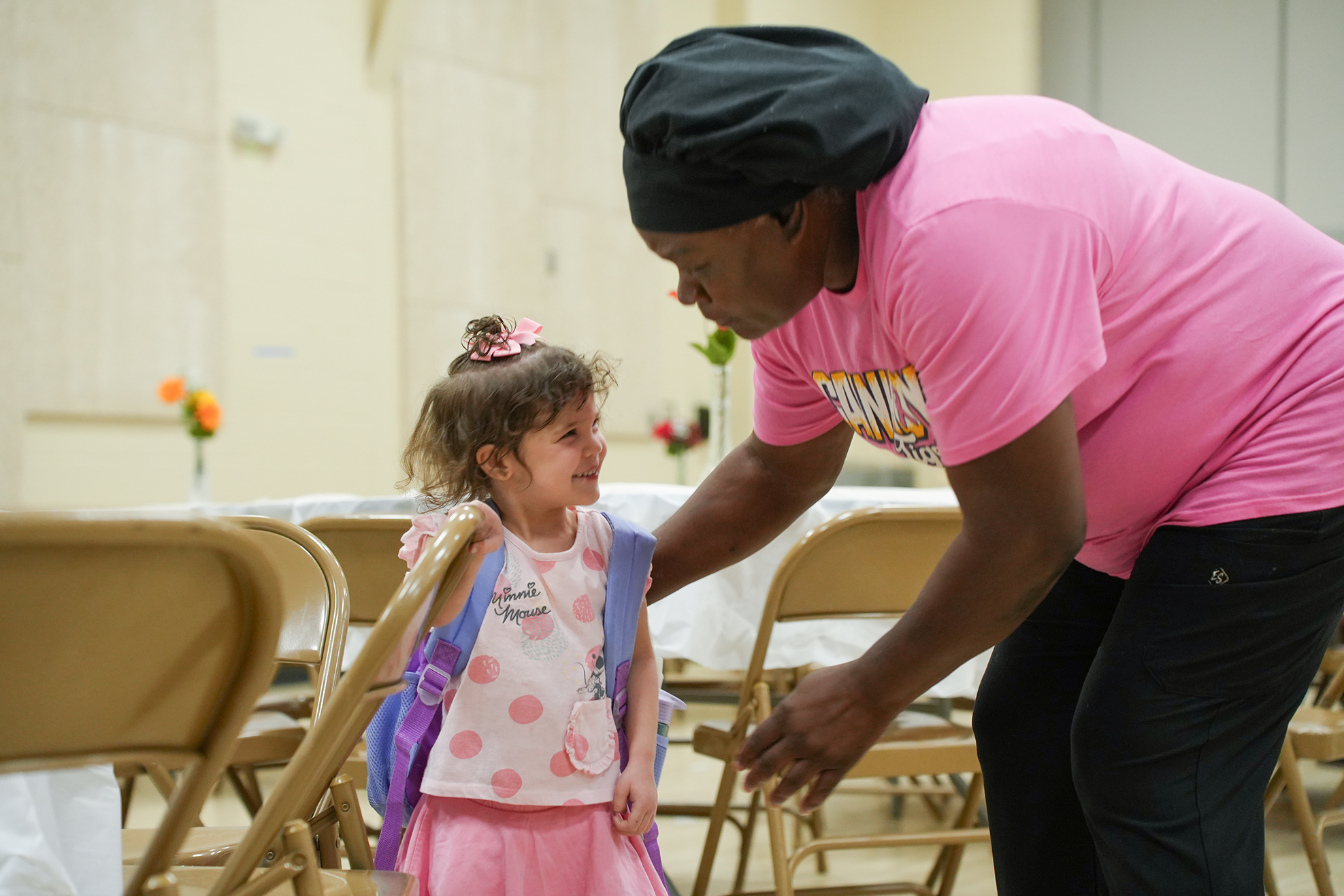 A woman in a pink shirt helps a young girl with a backpack in a room with chairs.