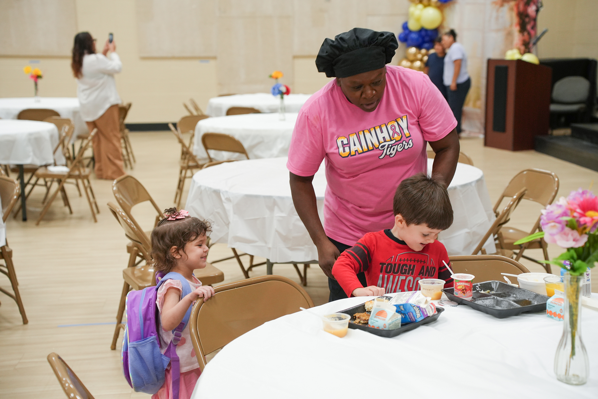 A woman in a pink shirt serves food to two children at a table, with other tables and chairs in a room.