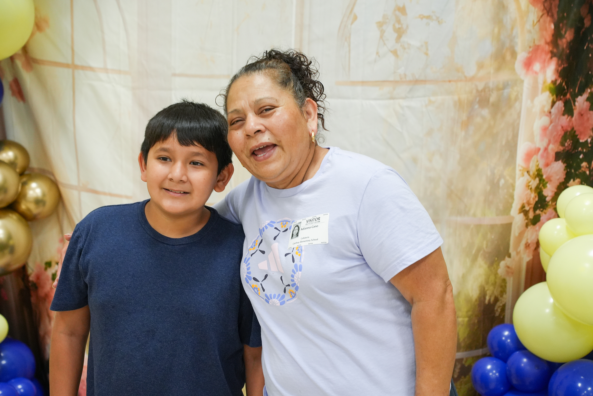 A woman and a young boy stand close together, smiling at the camera. Behind them, colorful balloons and a decorative backdrop.