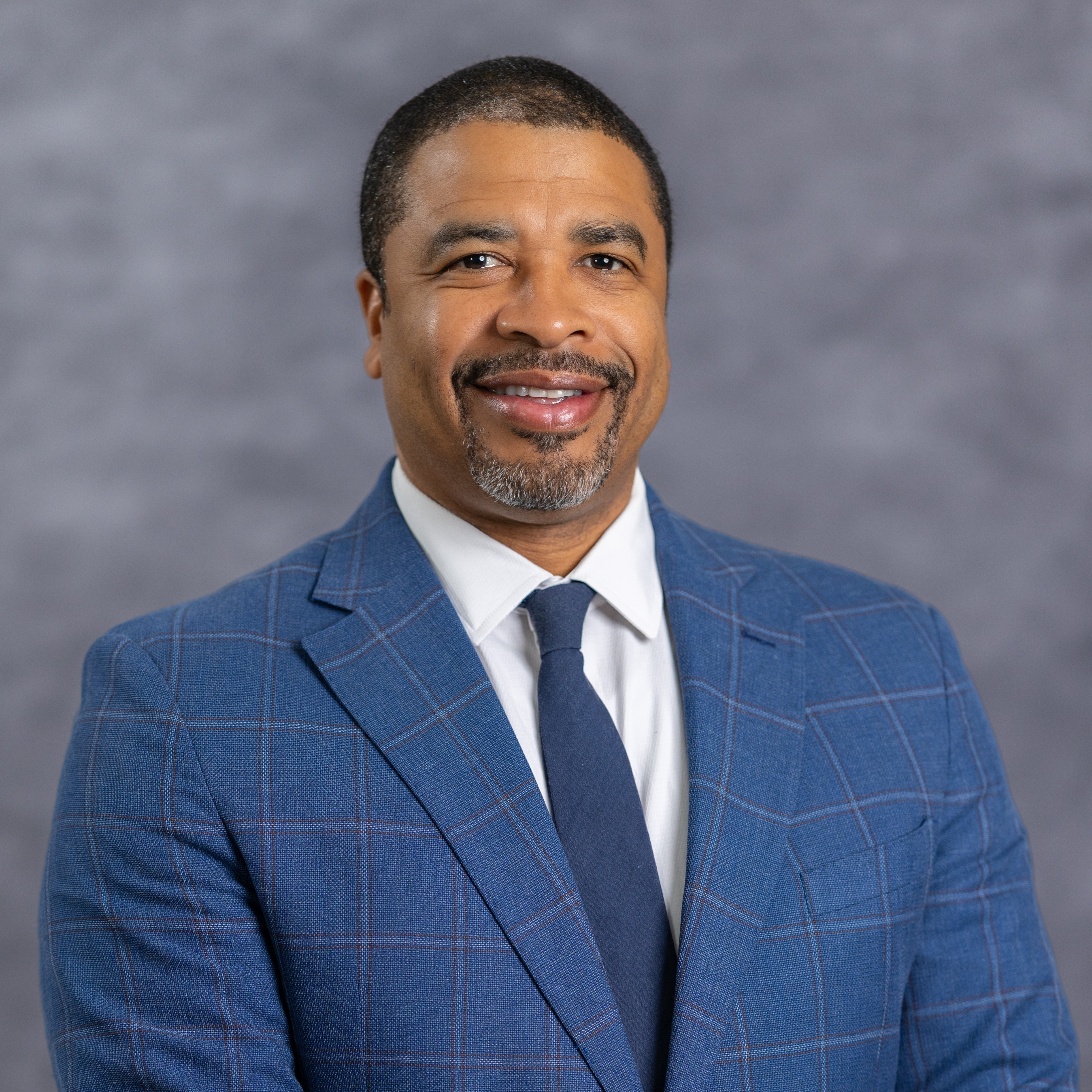 A man with a beard and short hair, smiling, wearing a blue suit with a white shirt and blue tie.