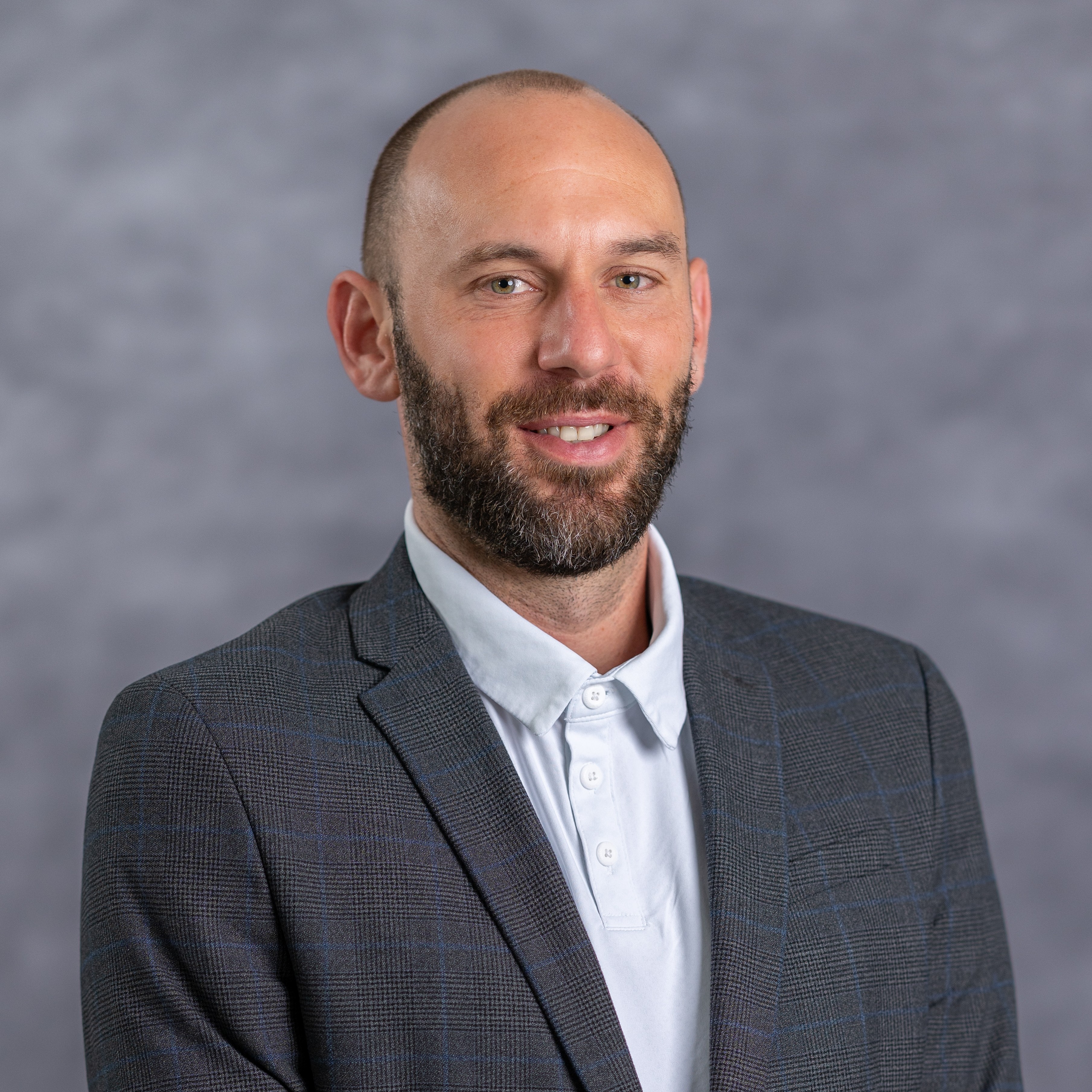 Man with beard in a light blue shirt and gray suit jacket smiles against a gray background.