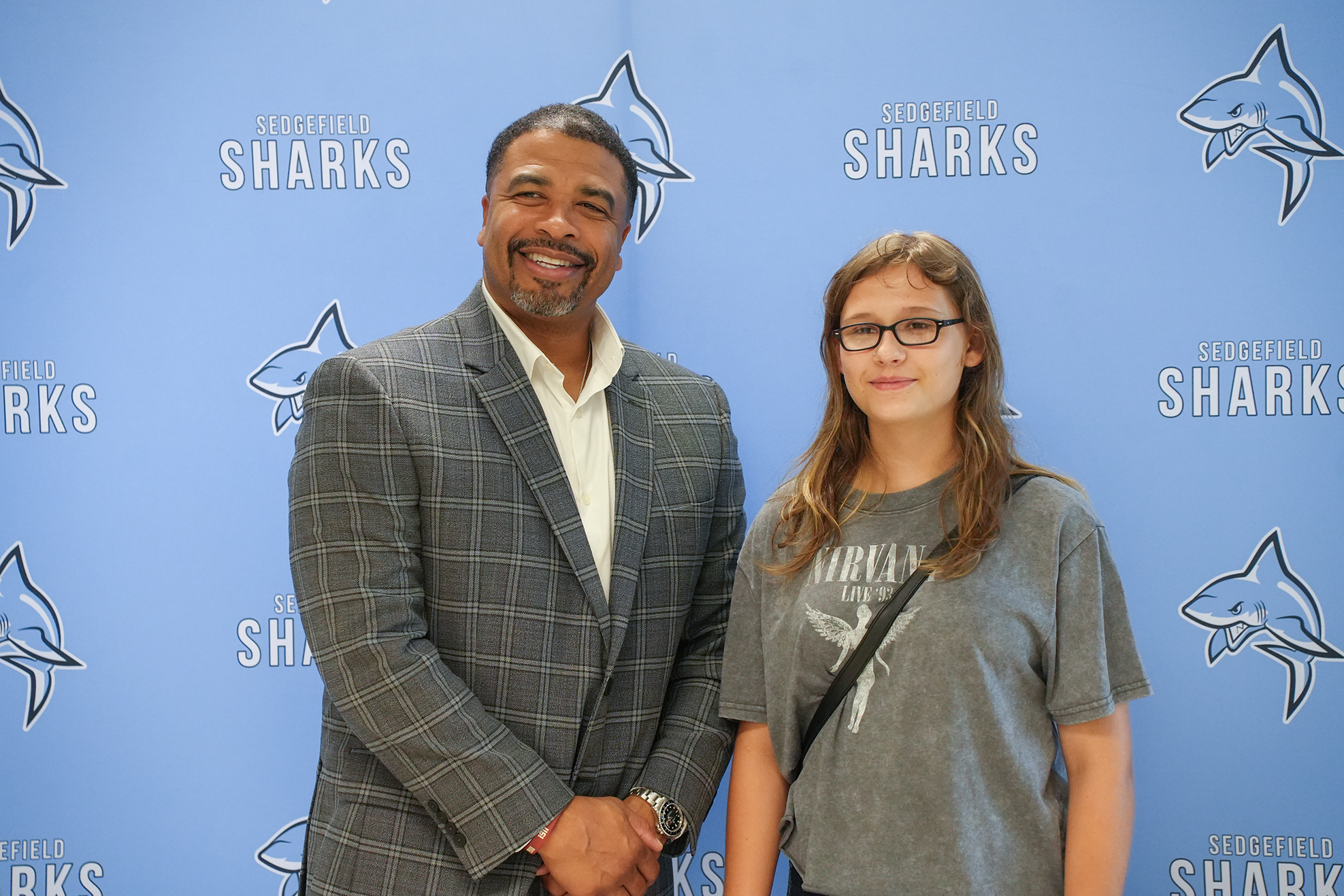 A man and a girl stand next to each other, smiling. Behind them is a blue wall with the word "Sharks" and shark images.