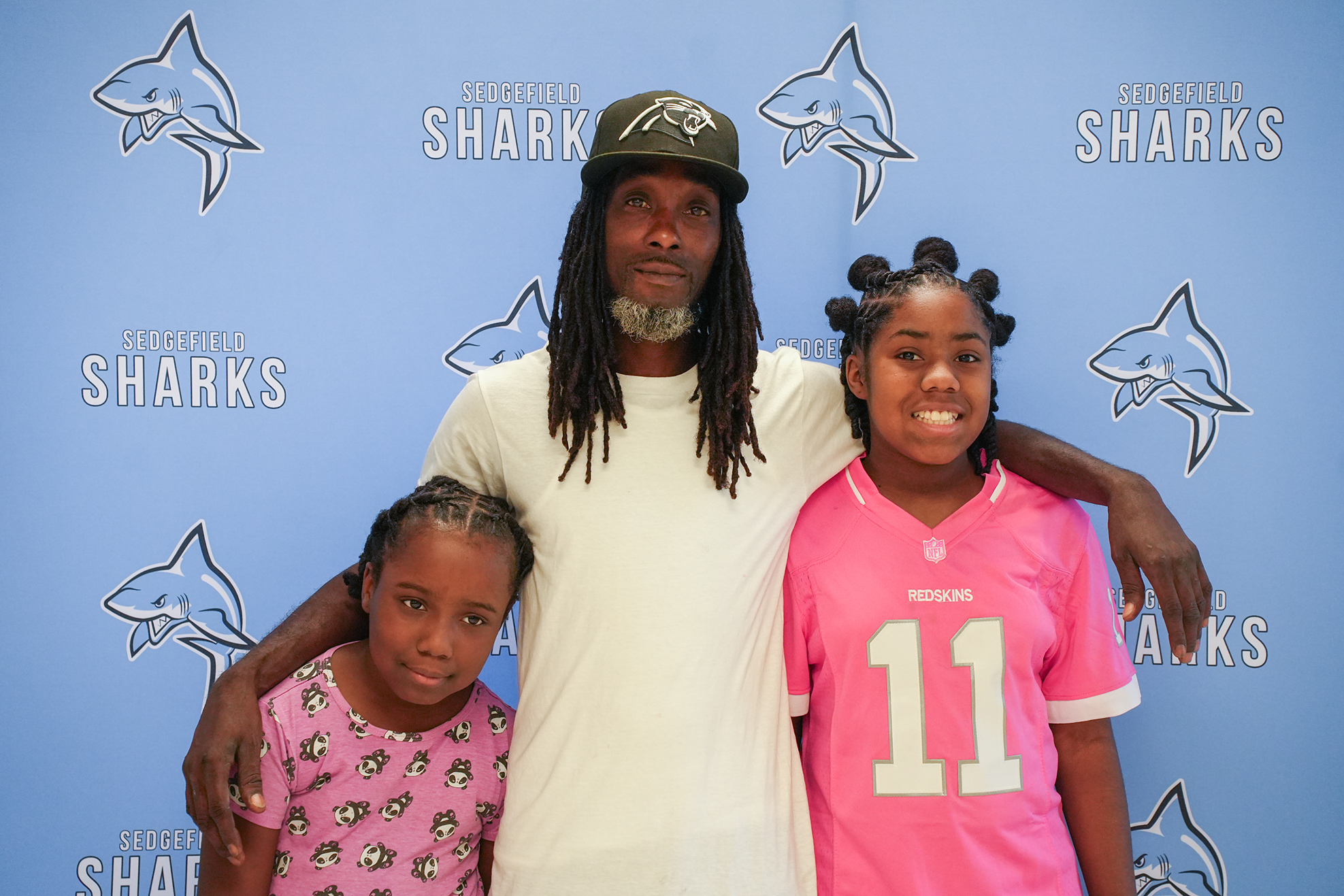 Three individuals pose for a photo in front of a blue wall with "Sharks" logos. The man has long dreadlocks and a cap, and his arm is around the two girls.