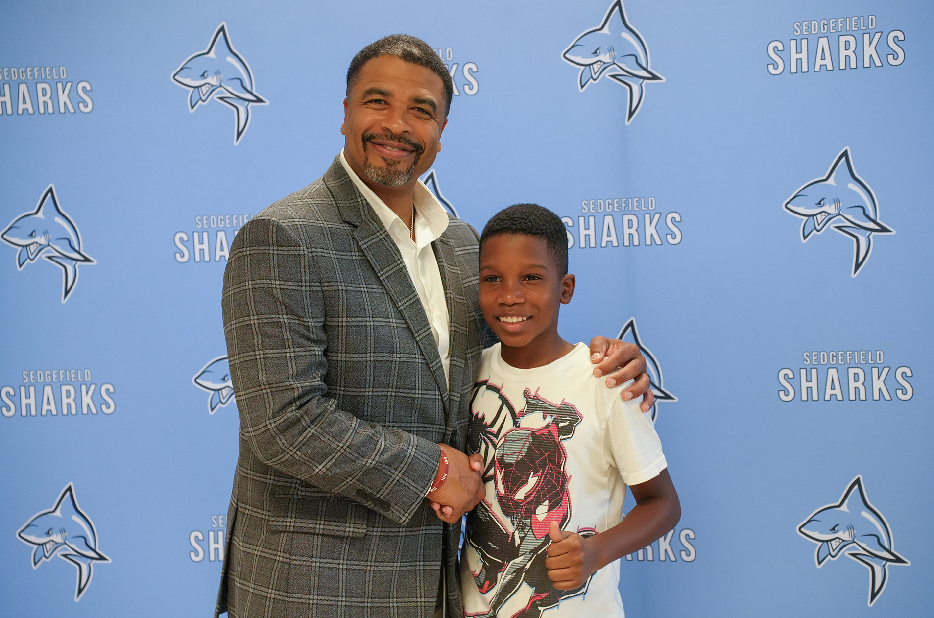 A man in a suit standing next to a boy holding a trophy, posing in front of a wall with shark logos.